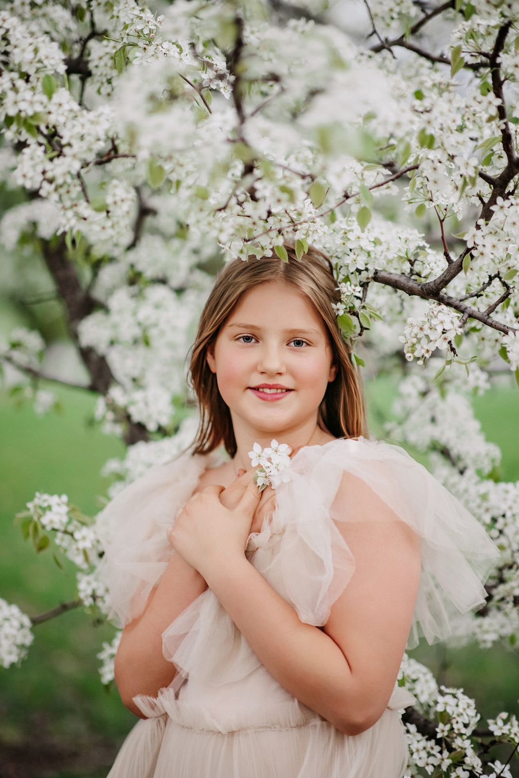 Young girl in blush tulle dress holding white flowers, posing before a blooming spring tree.