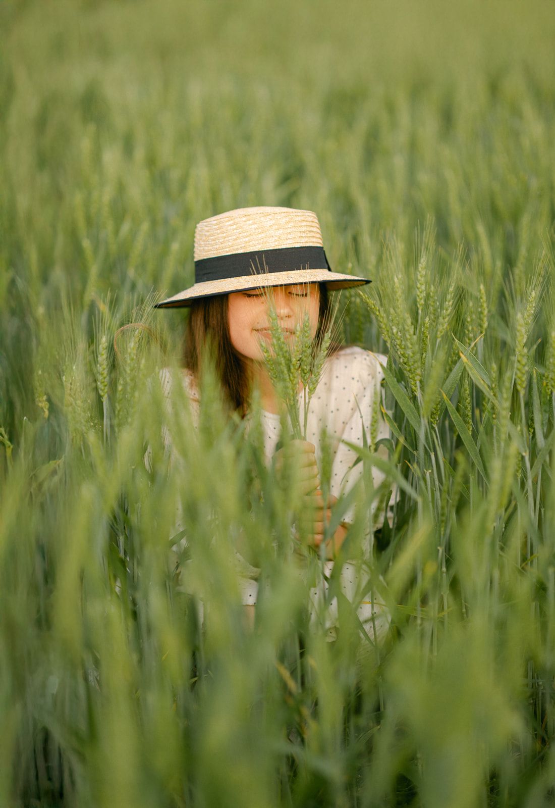 A young woman wearing a straw hat stands peacefully among tall green wheat stalks in a lush field.
