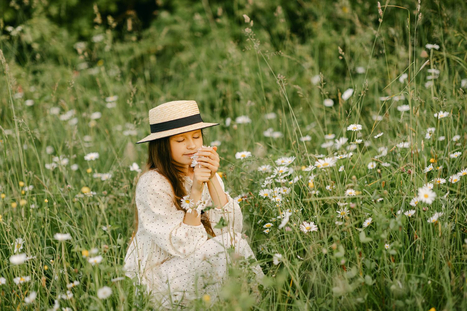 Young girl in white dress and straw hat holds flowers with eyes closed in daisy field.