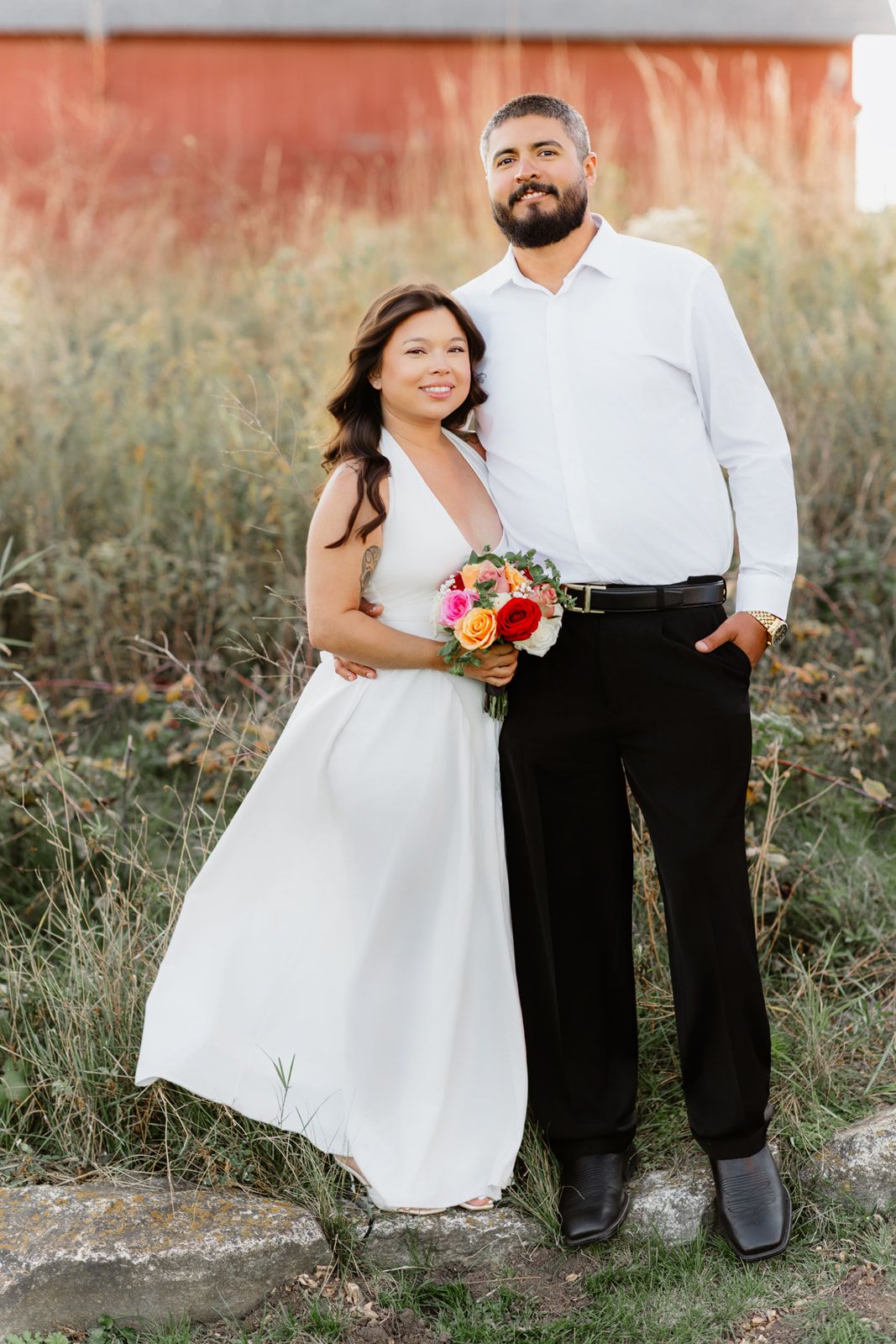Wedding couple posing outdoors, bride in white dress holding vibrant bouquet beside groom.