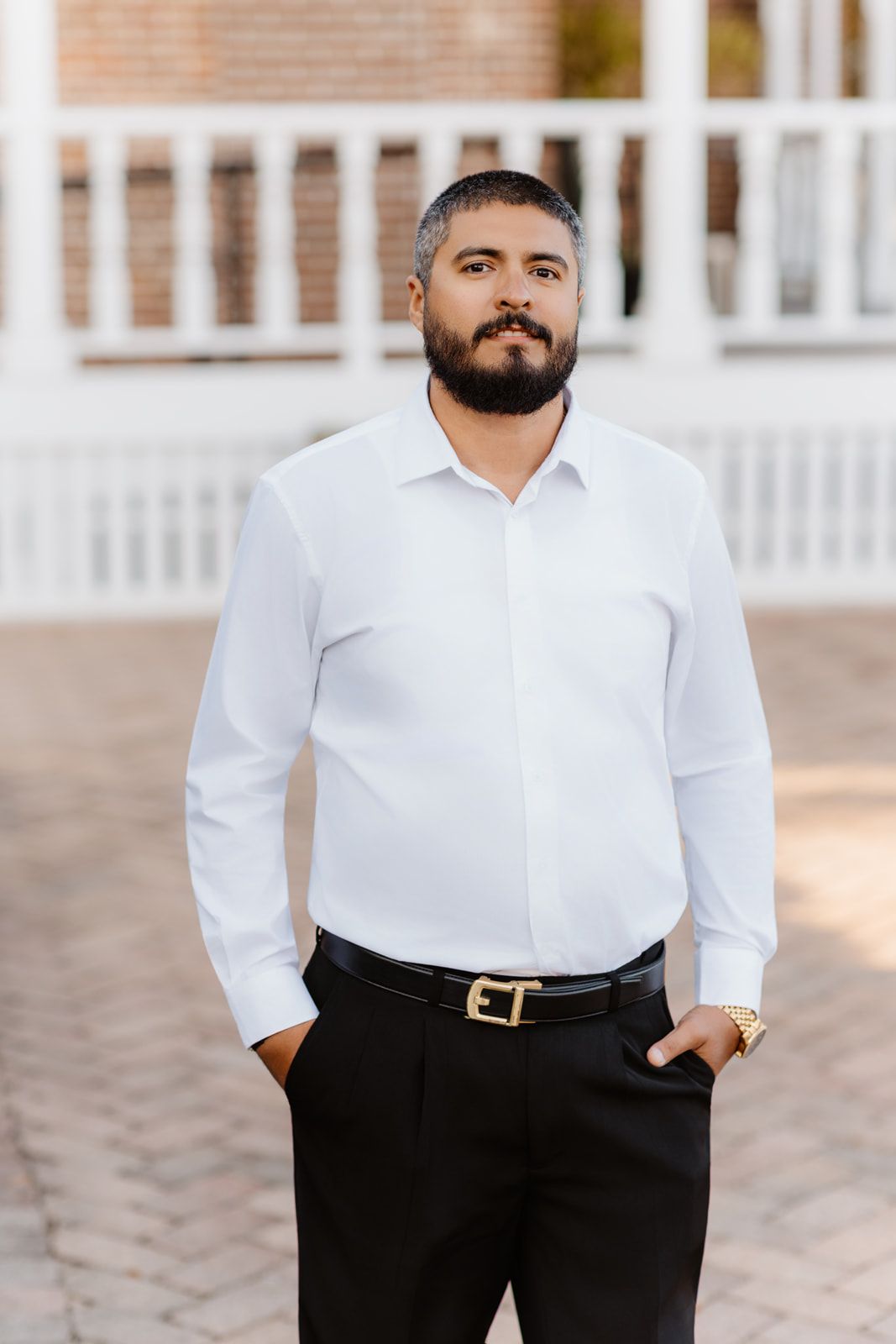 Handsome bearded groom in white dress shirt and black pants poses outdoors on brick pathway.