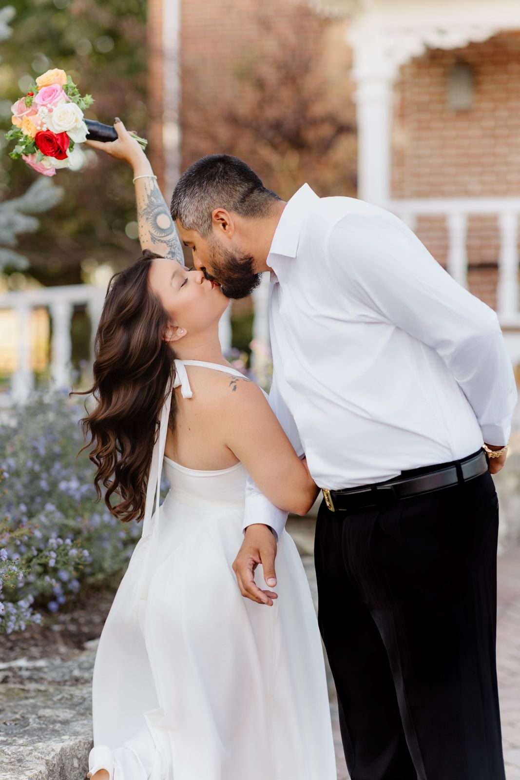 Bride and groom share a romantic kiss outdoors, bride holding colorful bouquet aloft in white dress.