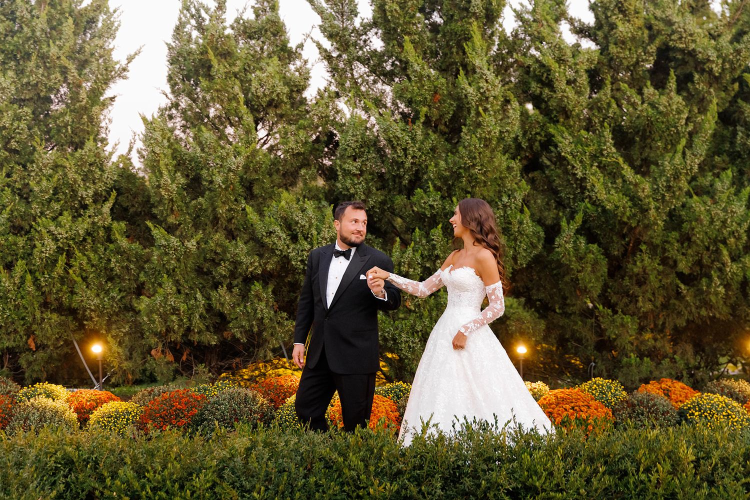 A bride and groom hold hands in a lush garden with colorful autumn flowers and tall evergreen trees at their wedding.