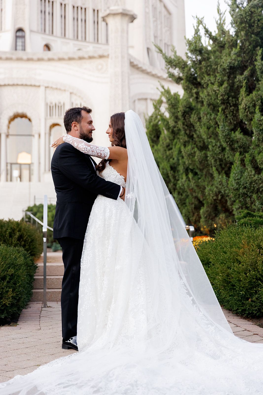 Bride and groom embrace outdoors near a grand building, her lace gown and long veil trailing elegantly on the ground.