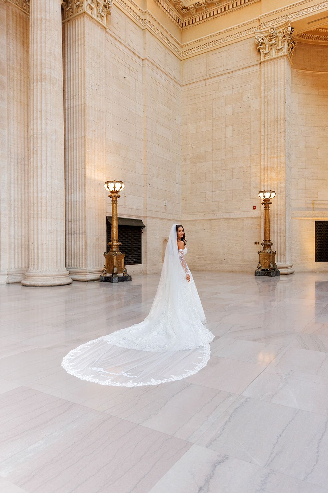 Bride in a white gown with a long cathedral veil stands in a grand marble hall with tall columns and gold accents.