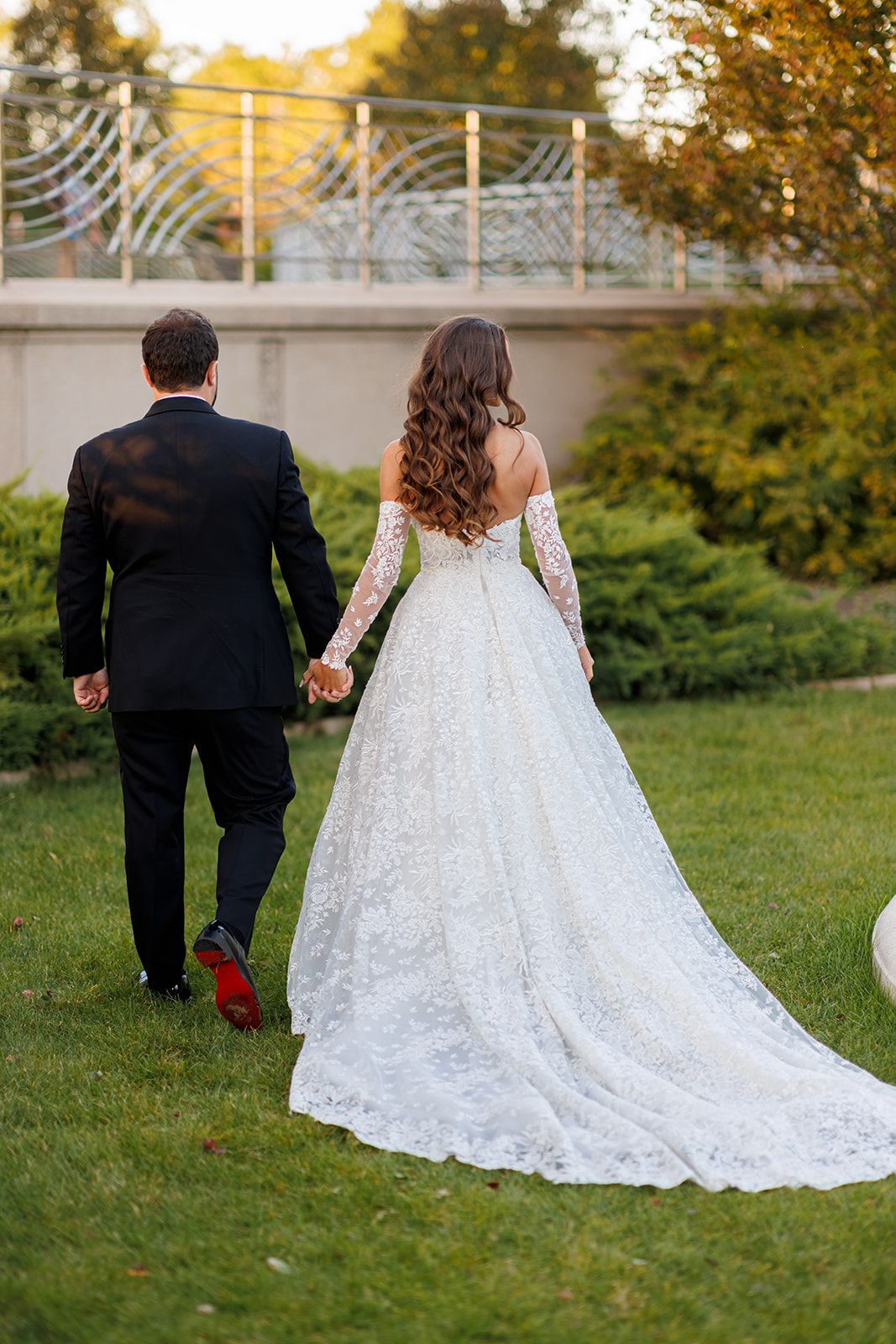 Bride in lace off-shoulder gown and groom in black suit walk hand-in-hand across green garden lawn.