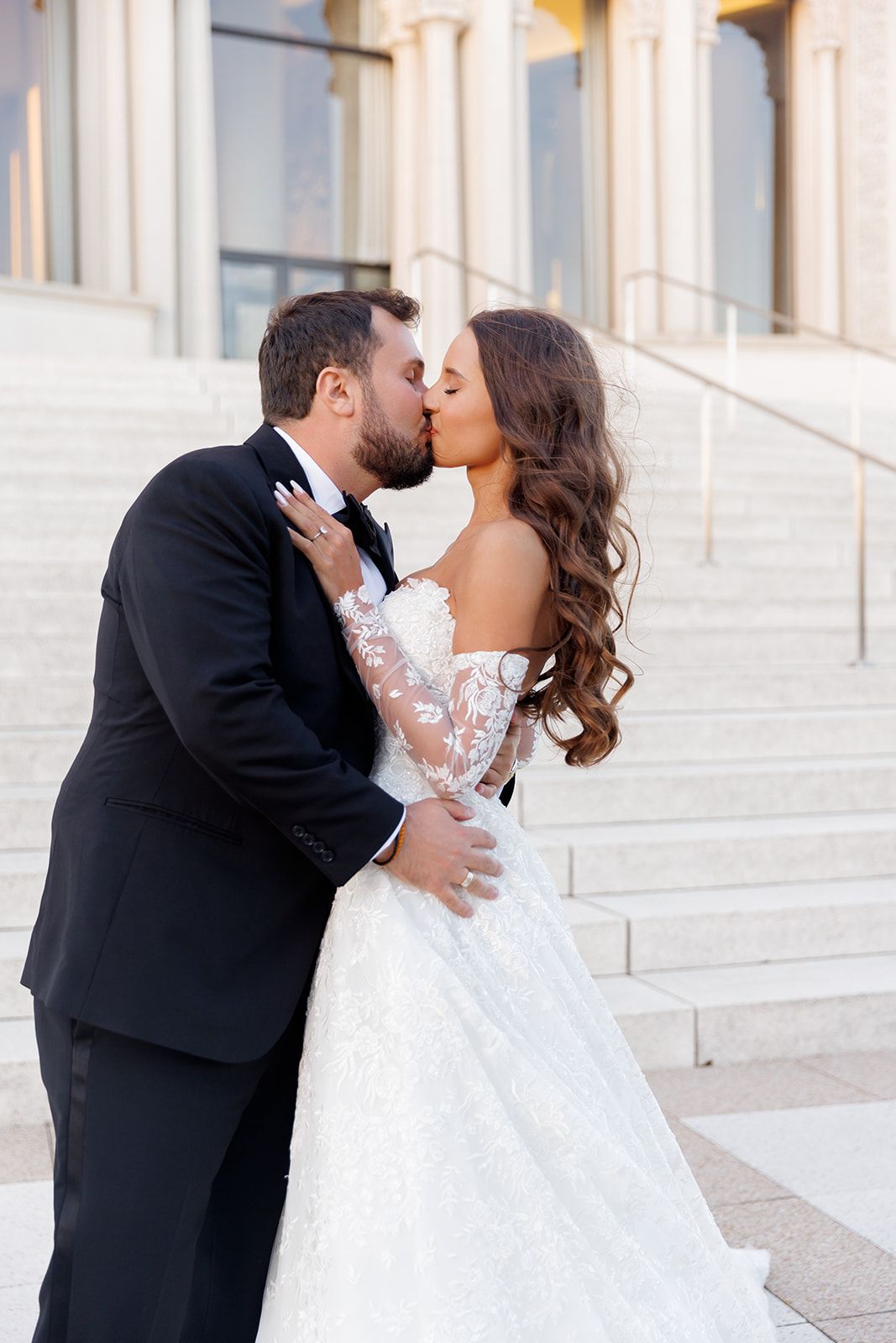Bride and groom share a romantic kiss on marble steps, bride wearing lace off-shoulder gown.
