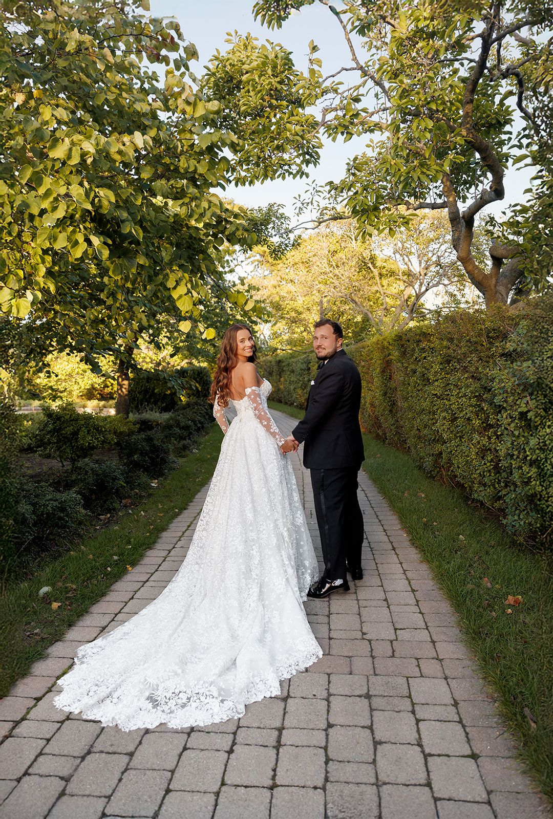 Bride in lace gown and groom in tuxedo hold hands on a garden pathway surrounded by lush green trees.