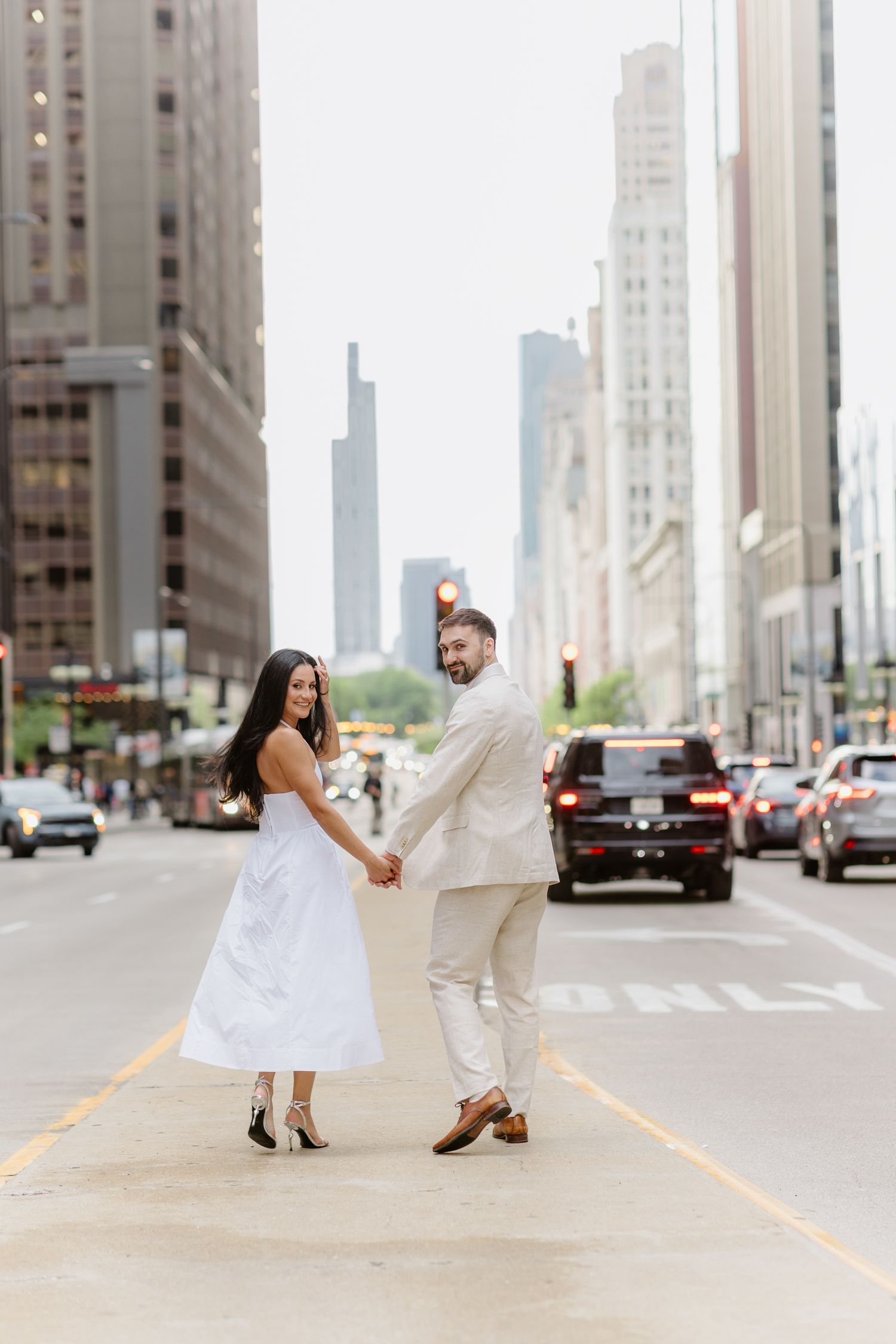 Couple holding hands in white outfits on a Chicago city street with skyscrapers in the background.