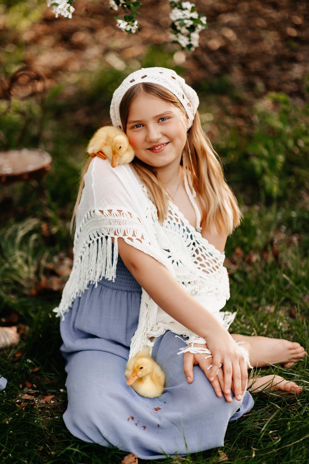 Young girl in white crochet top and blue skirt sits outdoors holding two yellow ducklings in a garden setting.