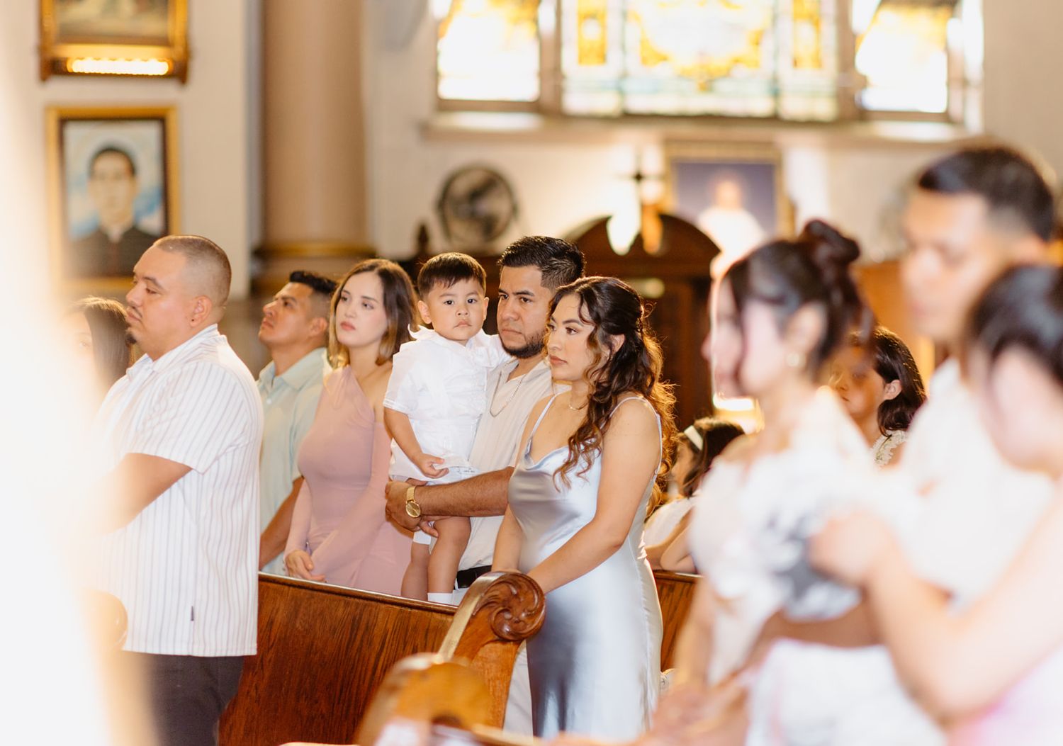 Family group holds a young child dressed in white during a baptism ceremony inside a Catholic church.
