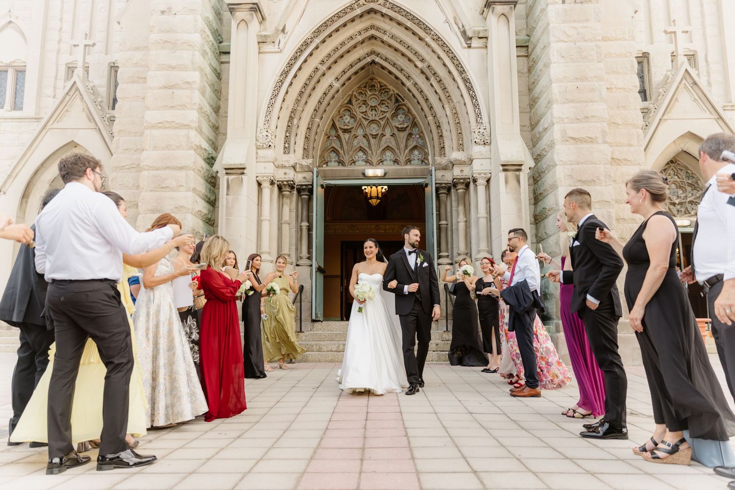 Newlyweds walk through cheering guests outside grand Gothic church after wedding ceremony.