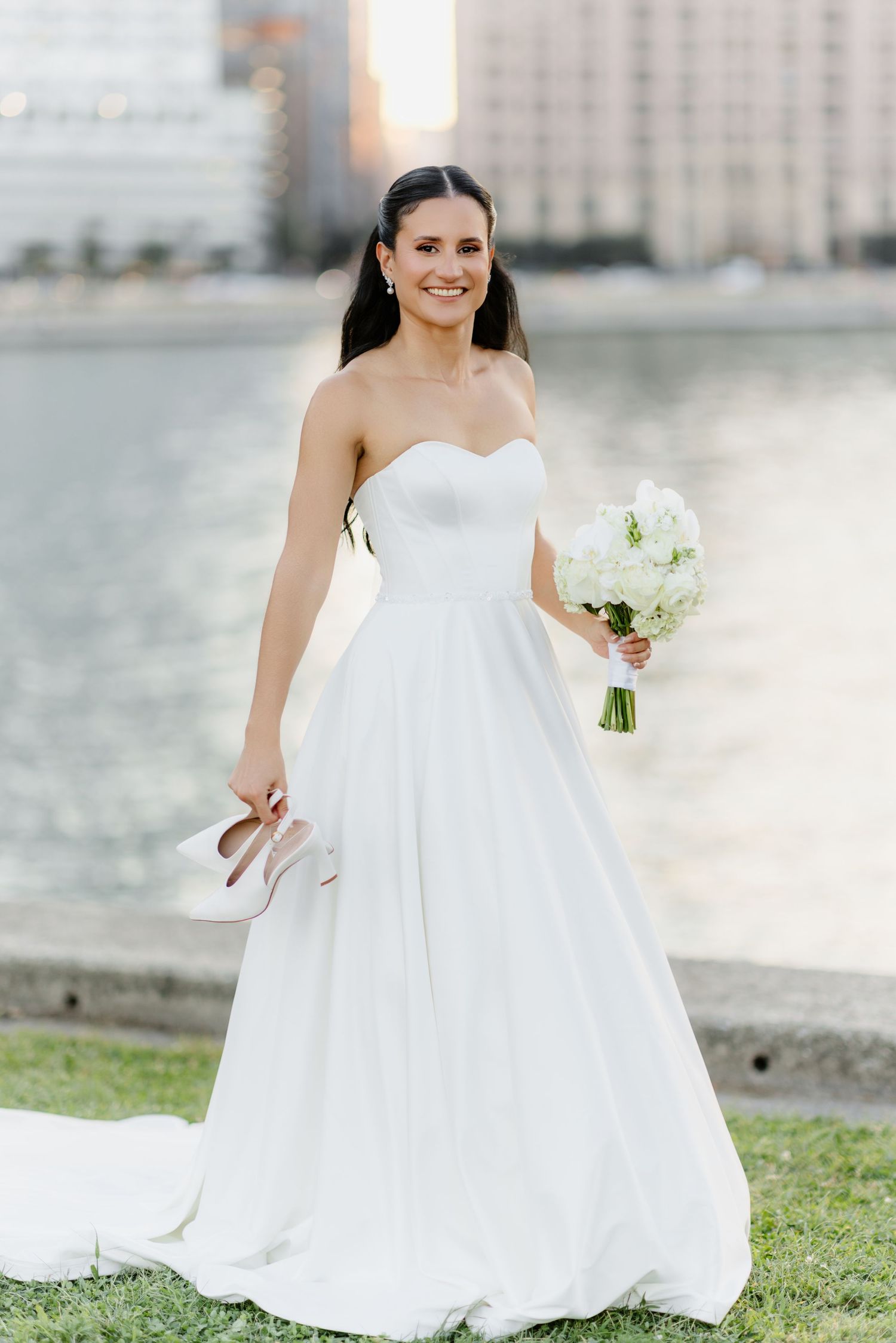 Joyful bride in strapless white gown raises bouquet triumphantly while holding heels by city riverfront.