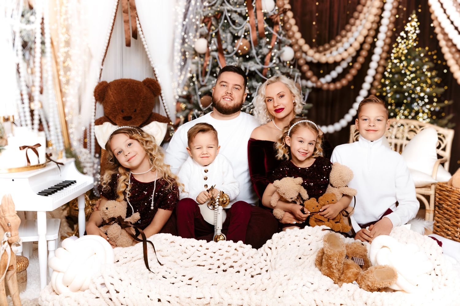 Family of five posing for a festive Christmas photo with teddy bears, holiday decorations, and a decorated tree.