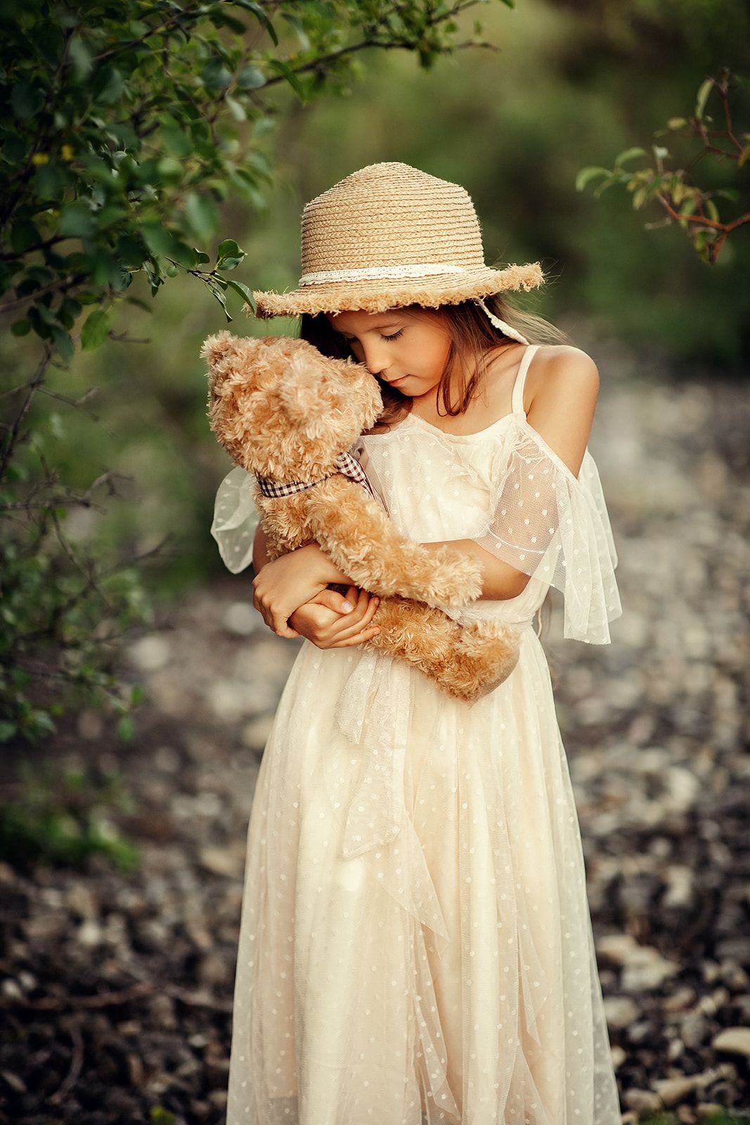 A young girl in a white dress and straw hat tenderly hugs a teddy bear outdoors in nature.