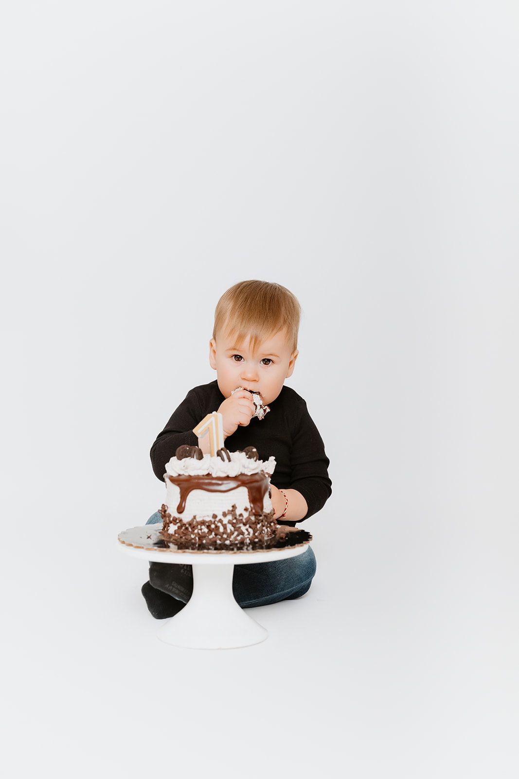 Baby boy in black sweater tasting first birthday chocolate cake on white background.