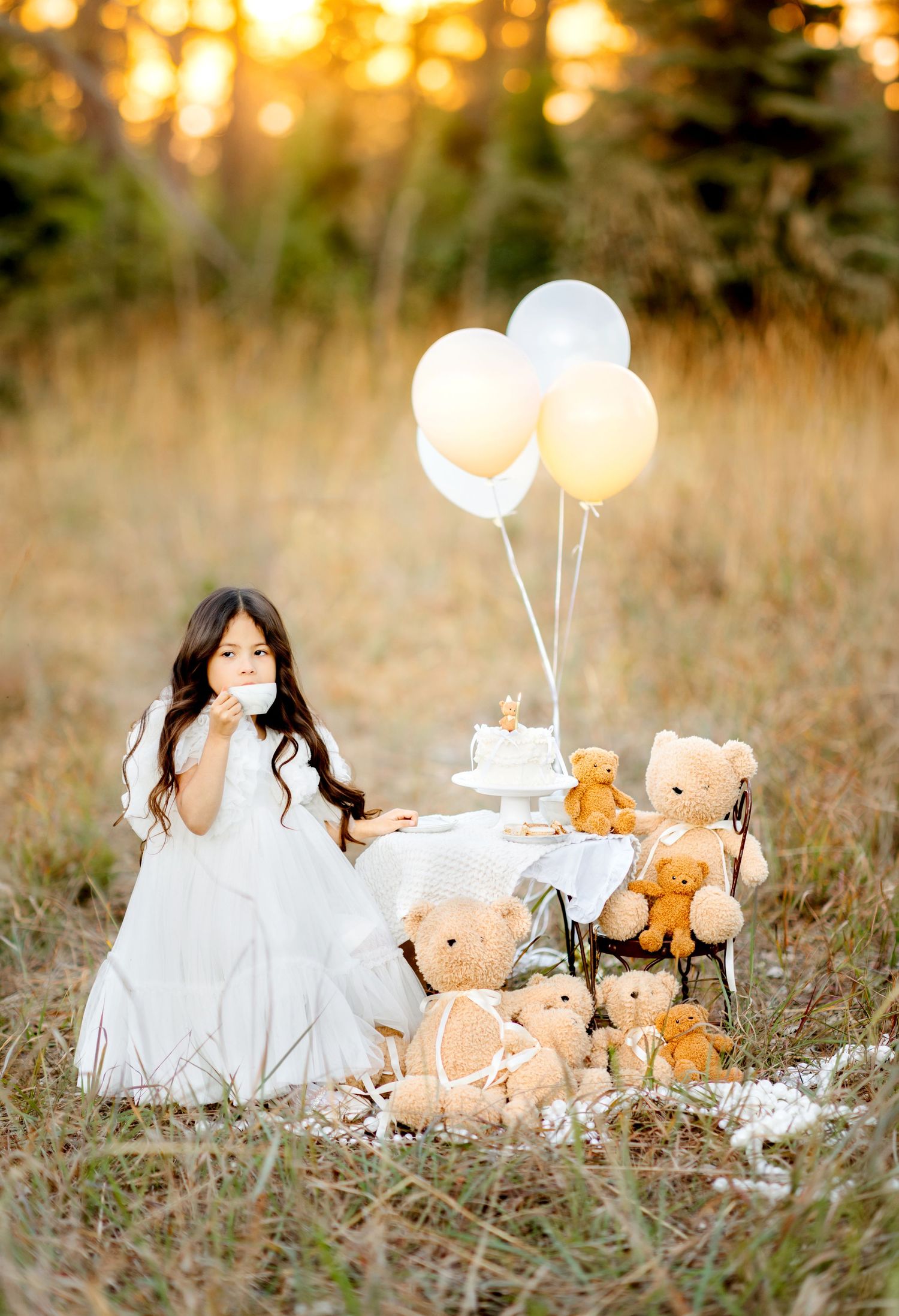 Girl in white dress at teddy bear tea party with balloons and cake in golden field at sunset.