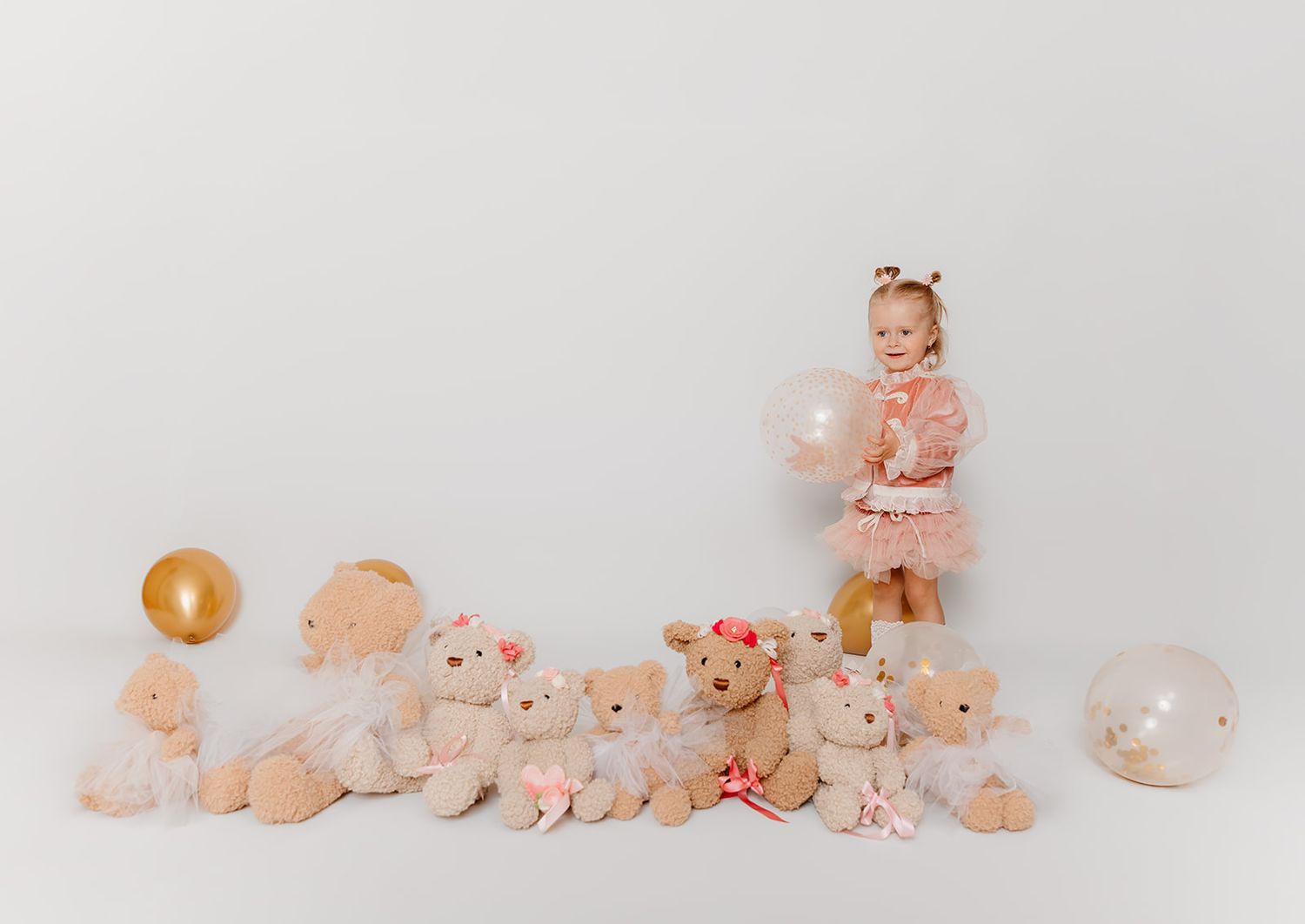 Toddler girl in pink tutu stands among teddy bears with balloons on white background.