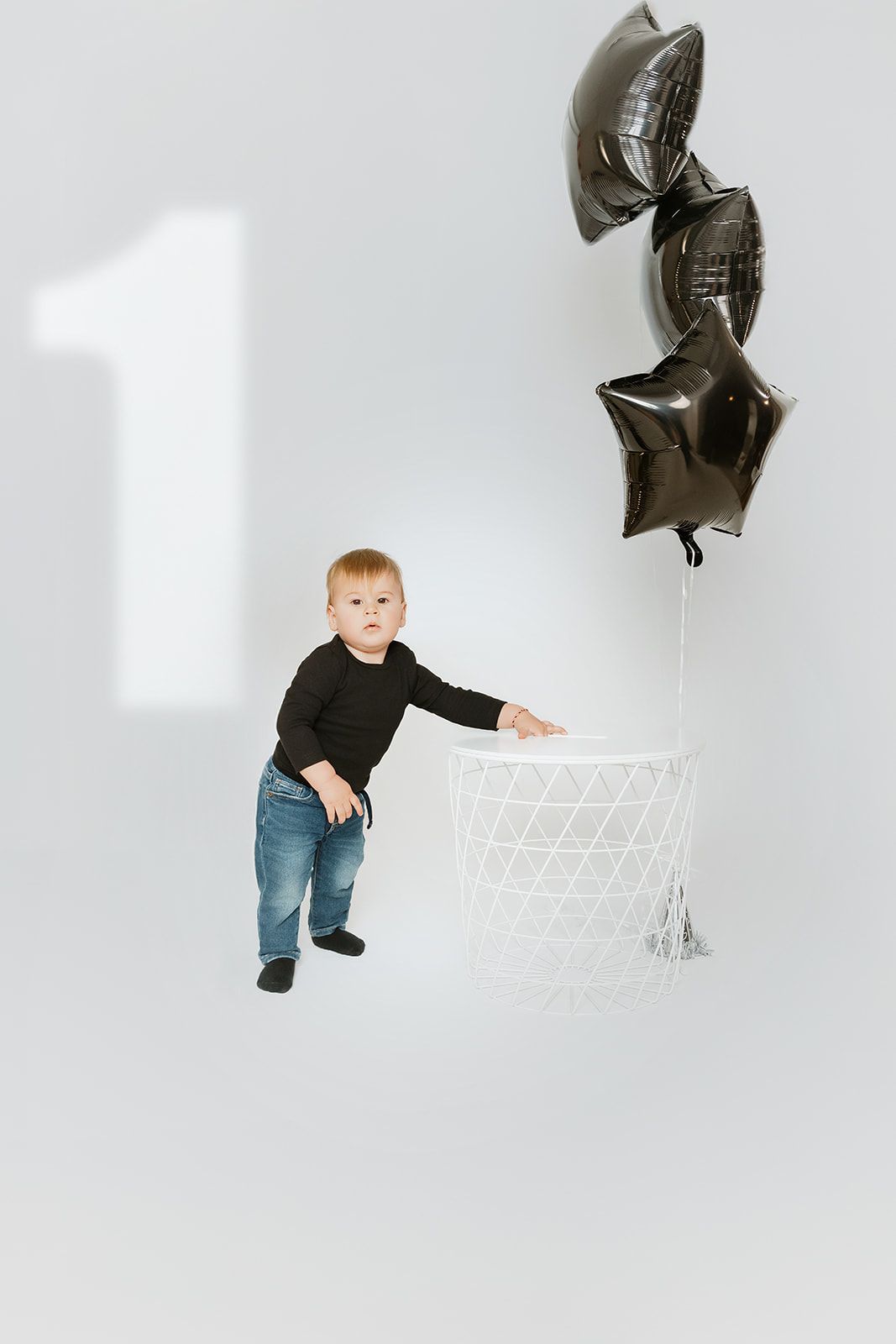 Toddler in black top and jeans stands by white basket with dark star balloons, celebrating first birthday.