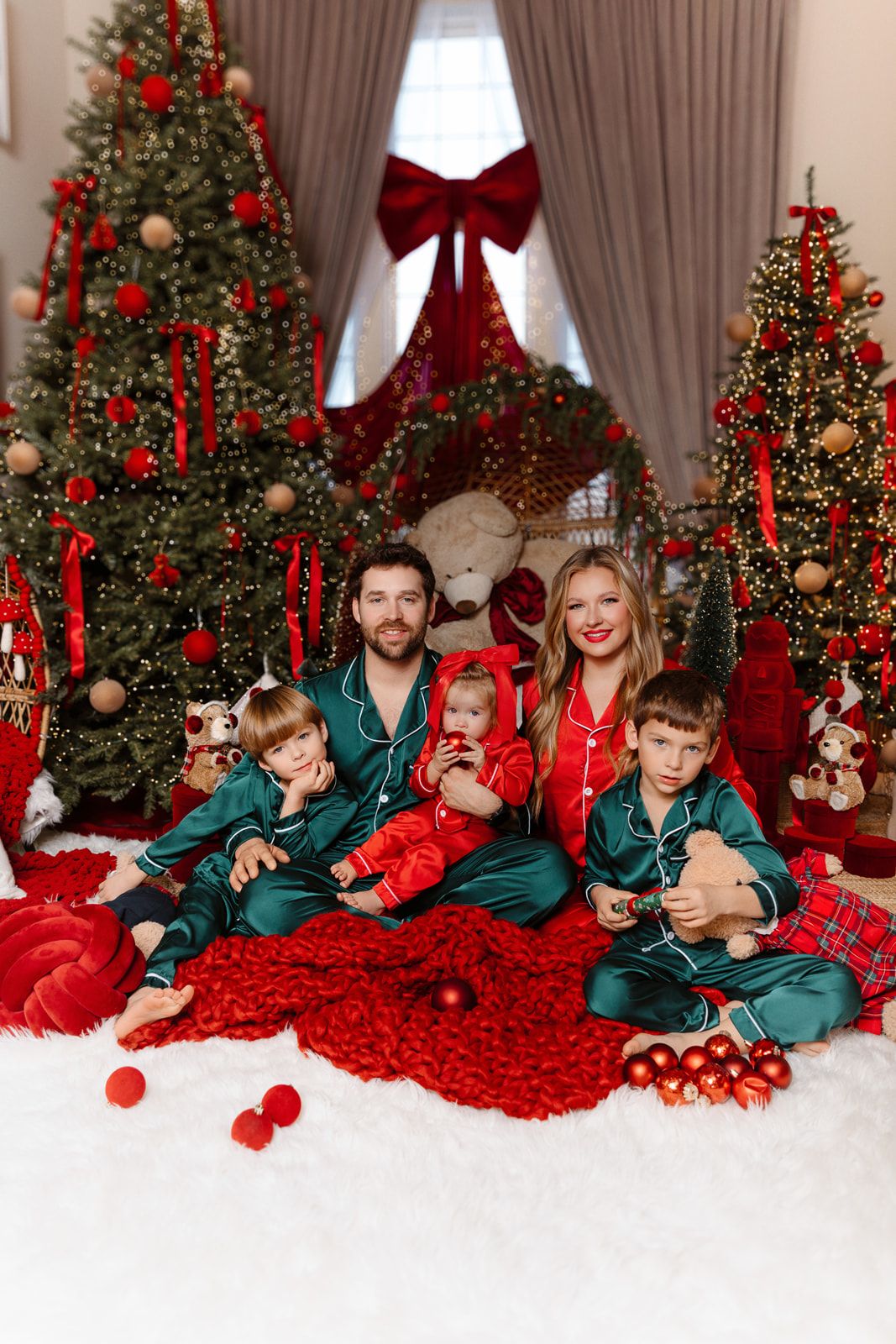 Family of five in green satin pajamas posing before decorated Christmas trees with red ornaments and holiday decor.