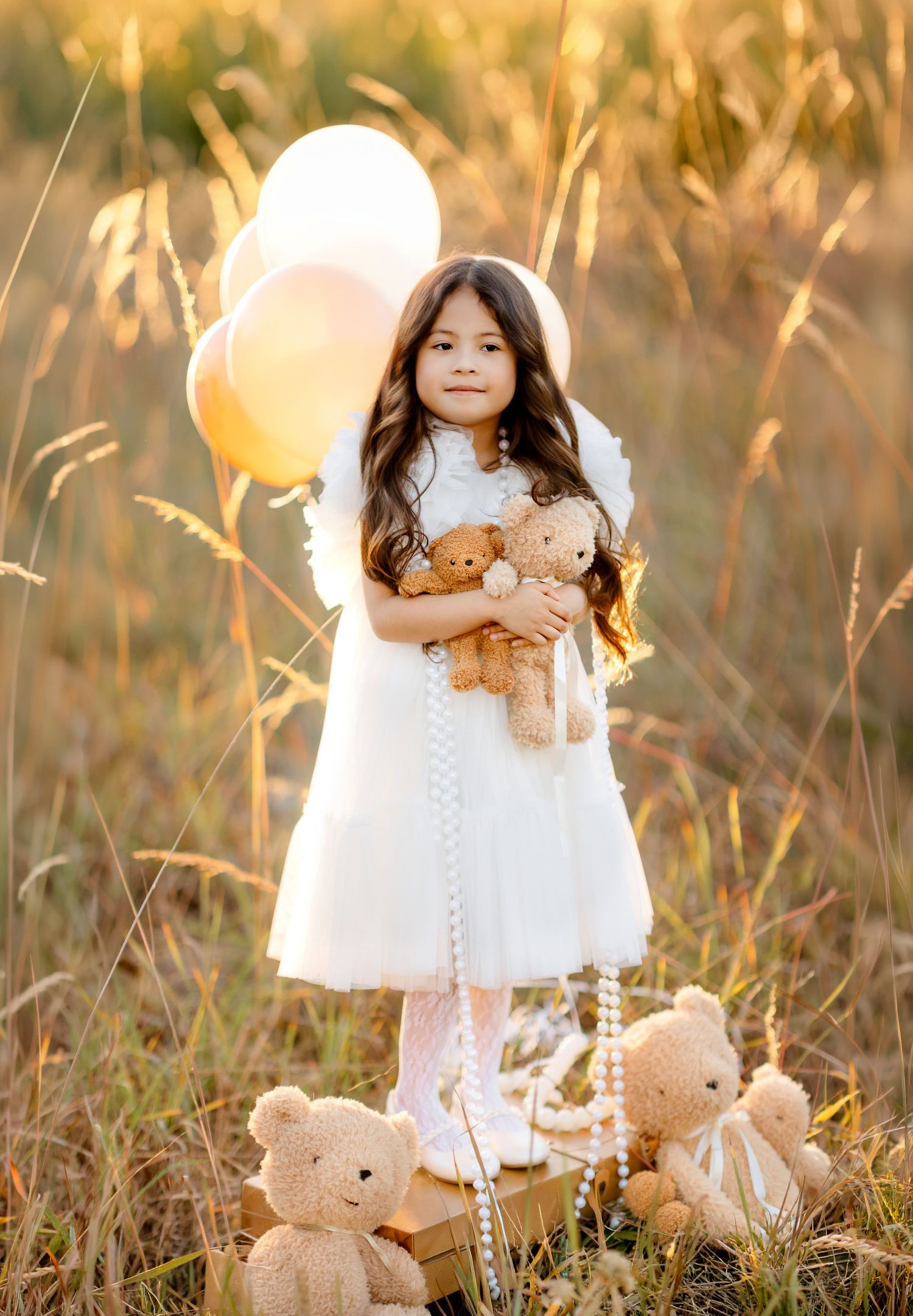 Young girl in white dress holding teddy bears with balloons in golden field at sunset.