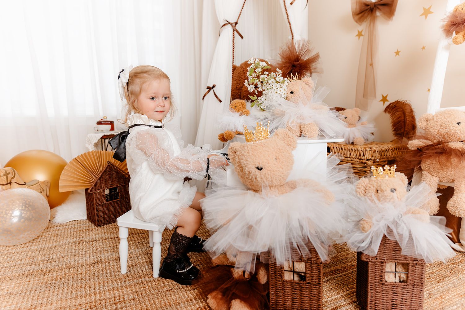 Toddler girl in white dress poses with teddy bear decorations and golden balloons in a boho birthday party setup.