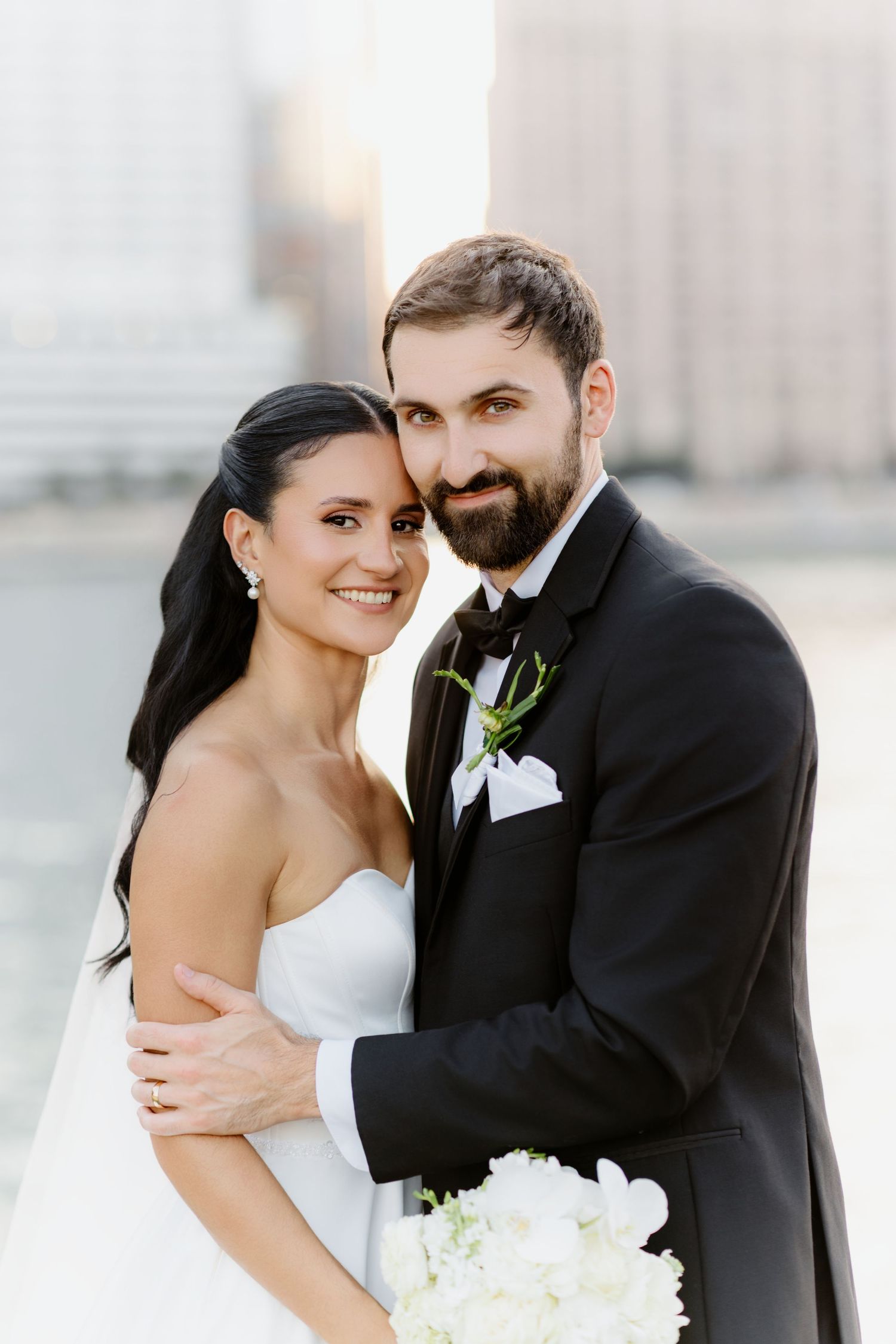 Bride and groom embrace closely, smiling at camera, she in strapless white gown, he in black tuxedo with bow tie.