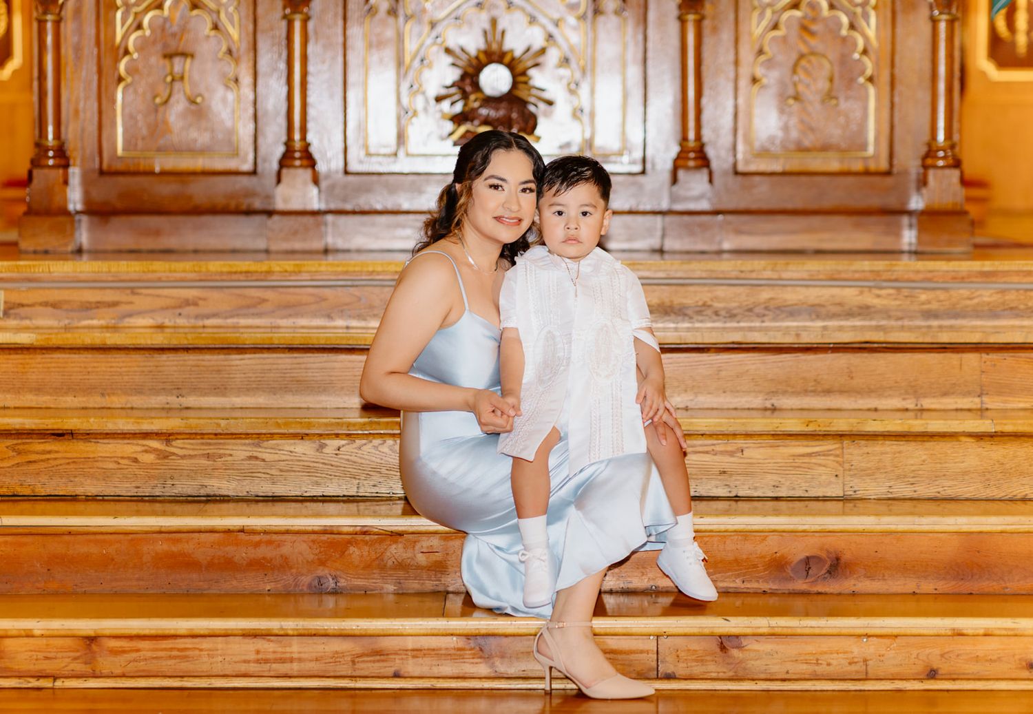 A woman in a light blue dress sits on ornate golden stairs holding a young boy in white baptism attire.