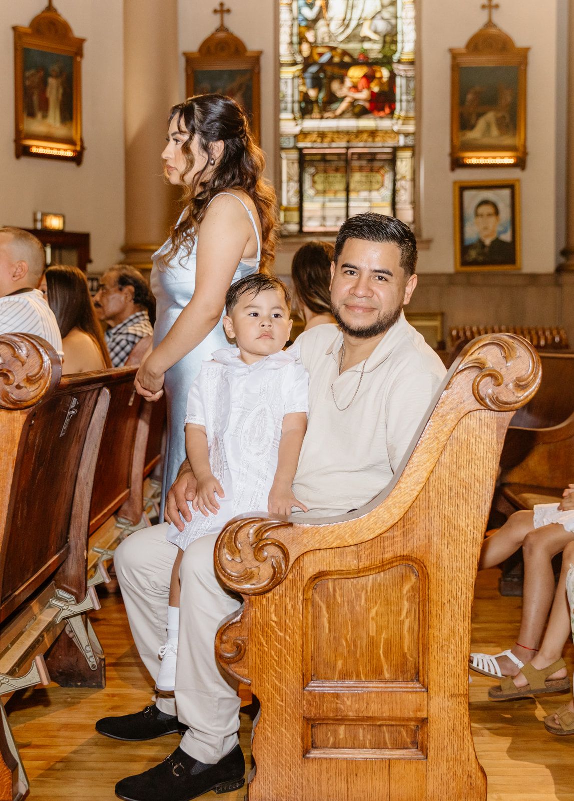 A family sits in wooden church pews during a baptism, with a young boy in white between his parents.
