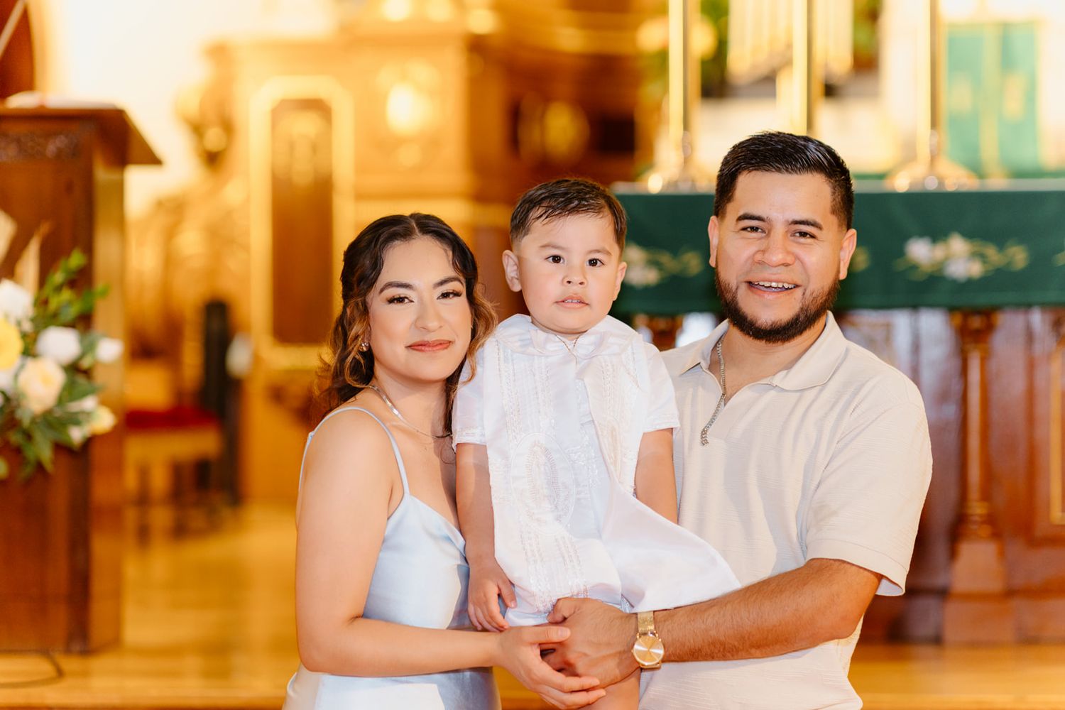 Family of three poses in church, parents holding baby dressed in white christening outfit.