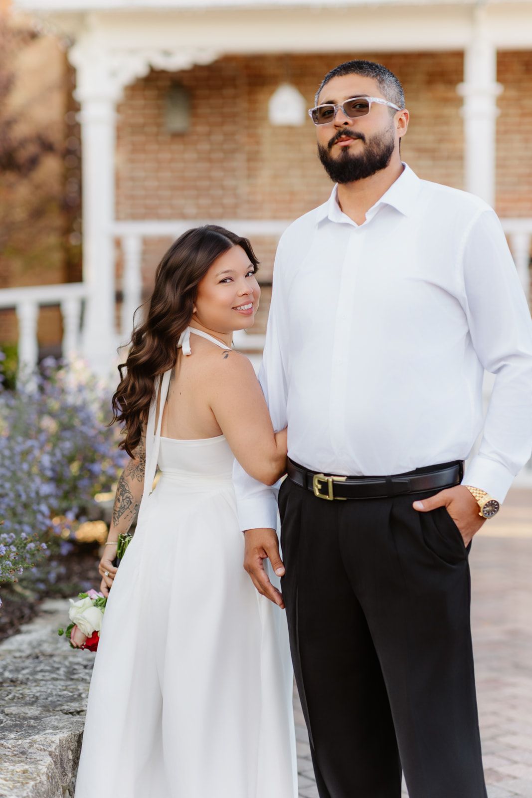 Bride in white halter dress and groom in white shirt posing together near a brick building with flowers.