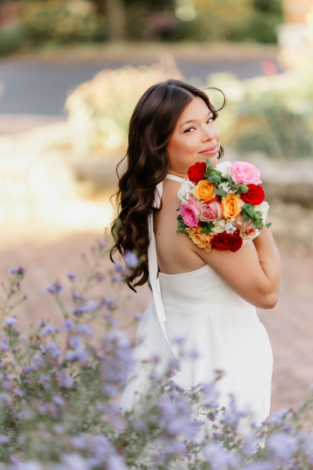 Bride in backless white dress holding vibrant rose bouquet, smiling over shoulder among purple flowers.