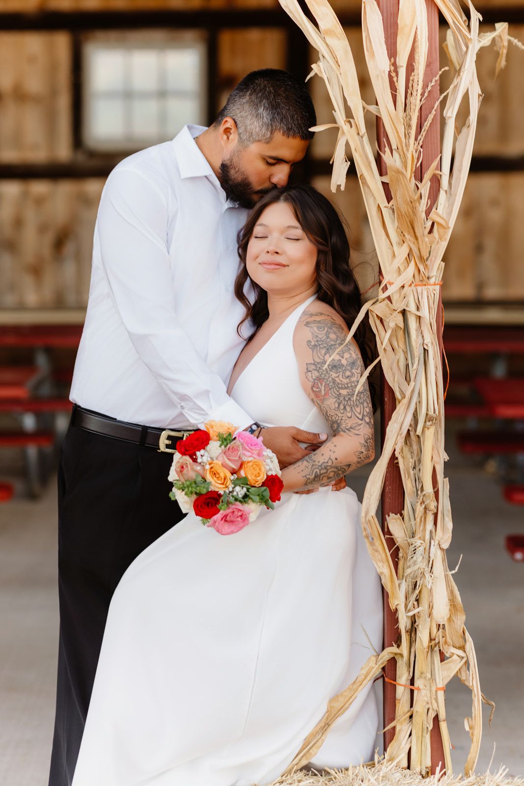 Bride and groom embrace near rustic barn decor, she holds colorful bouquet in white gown, he wears white dress shirt.