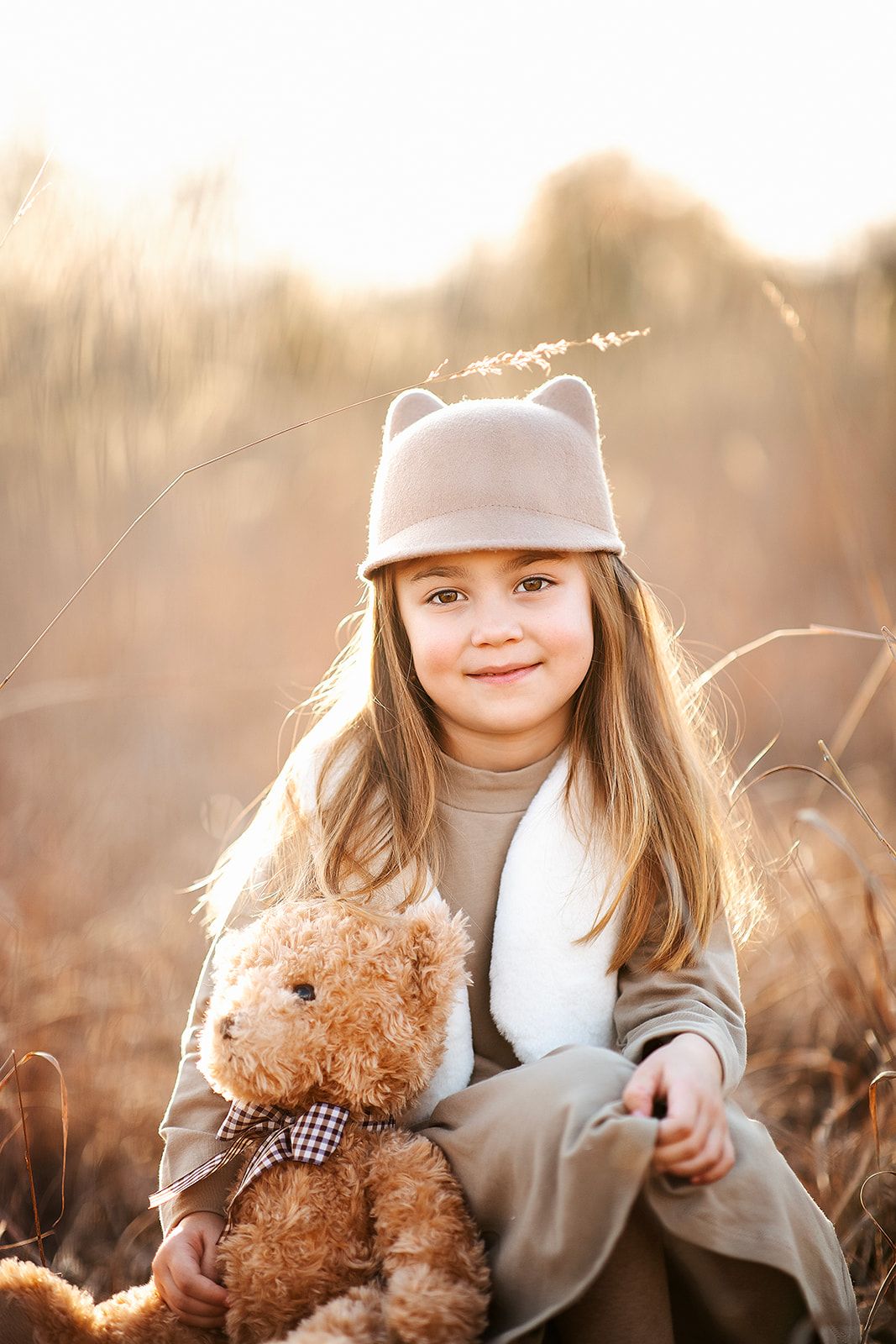 Young girl in cat-ear hat holds teddy bear in golden field at sunset, smiling warmly at camera.