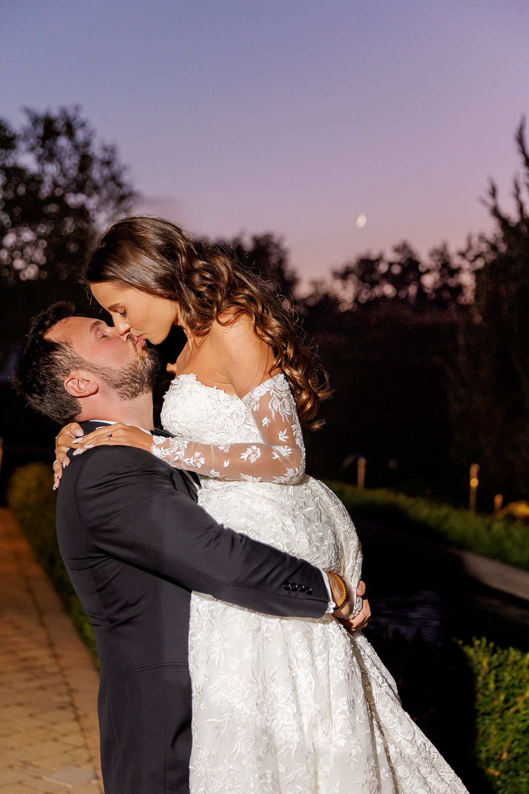 Groom lifts bride for a romantic kiss outdoors at dusk, with a crescent moon visible in the purple evening sky.