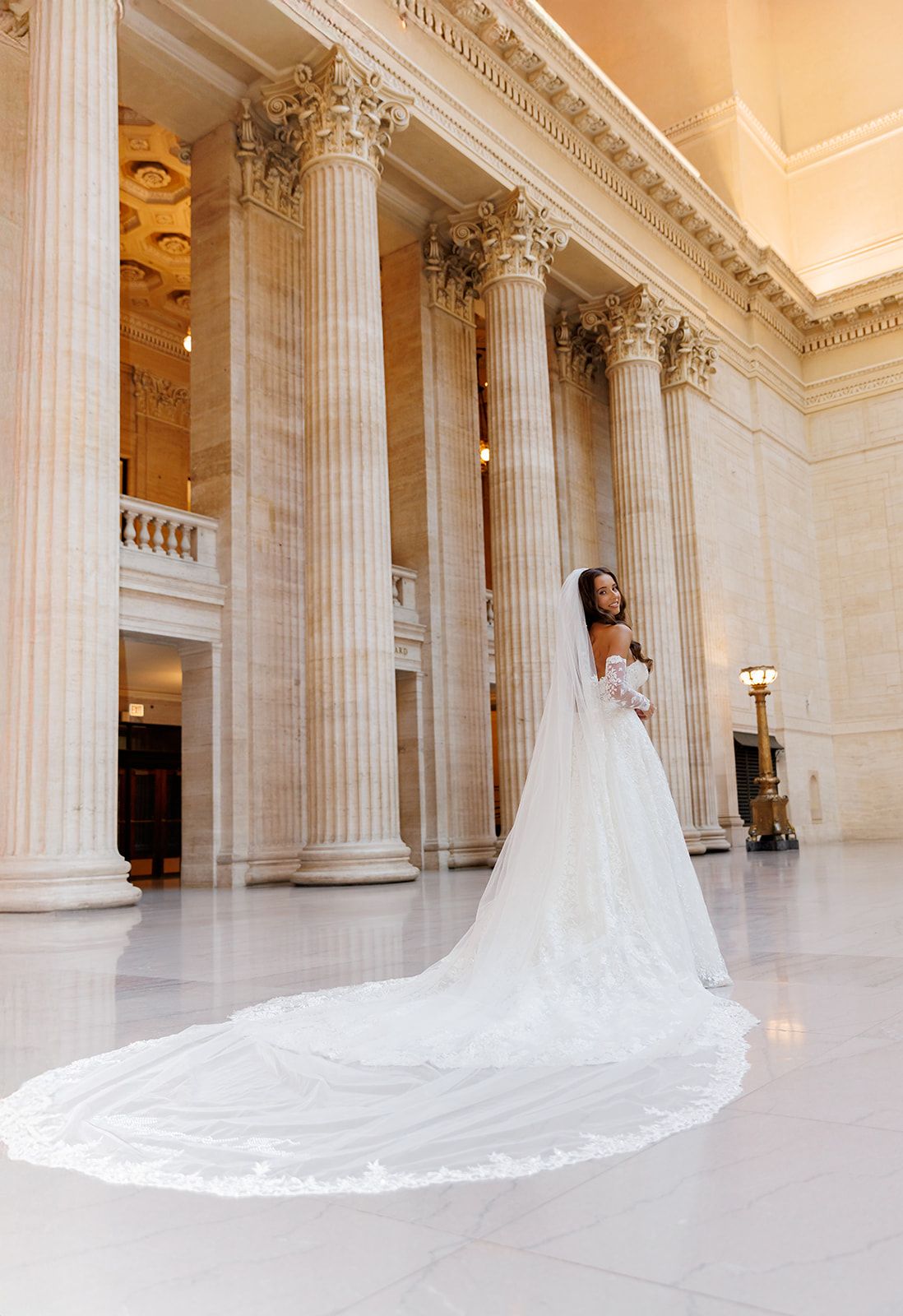 Bride in a stunning white ball gown with cathedral veil poses among grand marble columns in an elegant neoclassical hall.