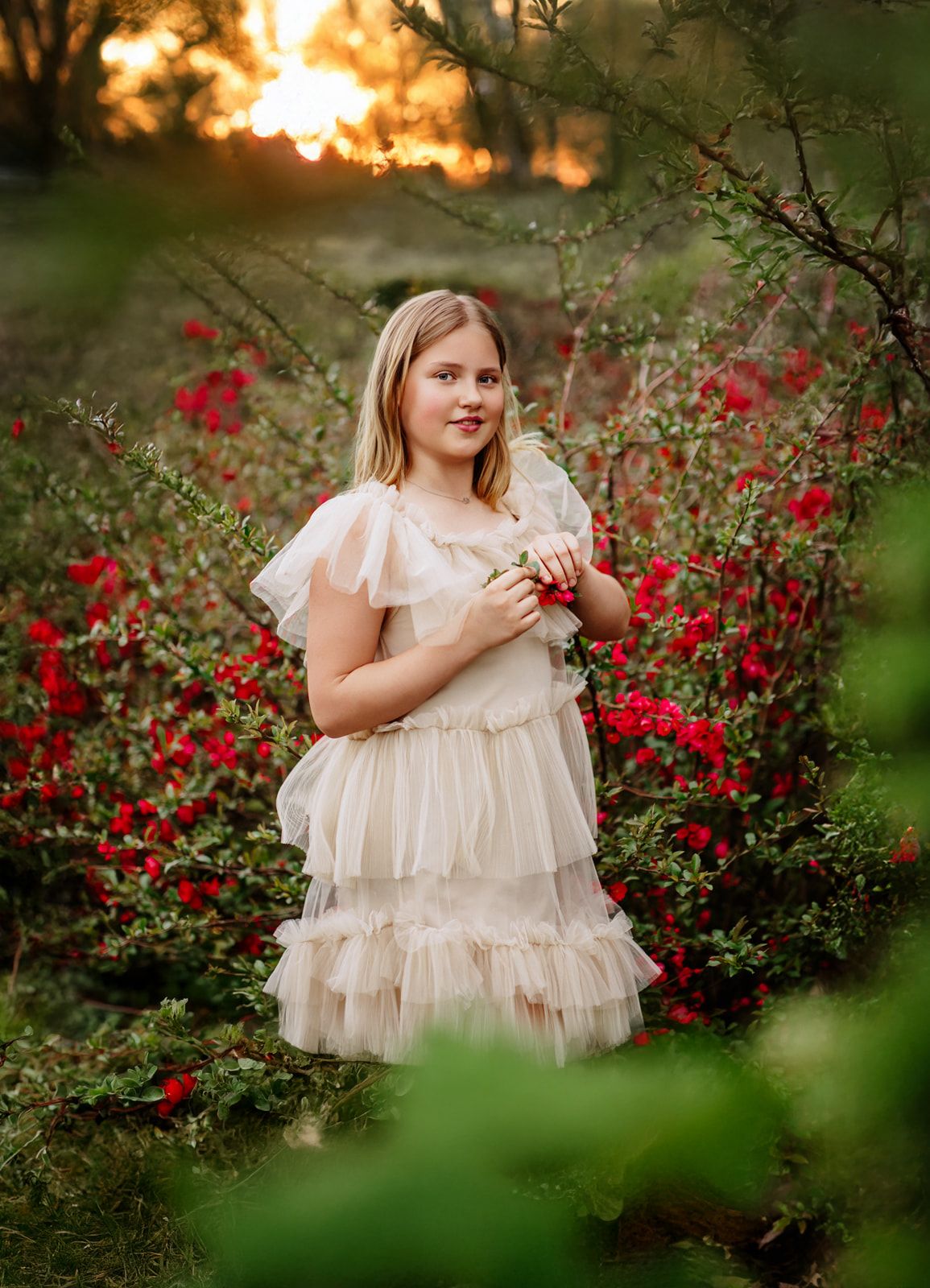 A young girl in a cream ruffled dress stands among red roses at golden hour sunset.