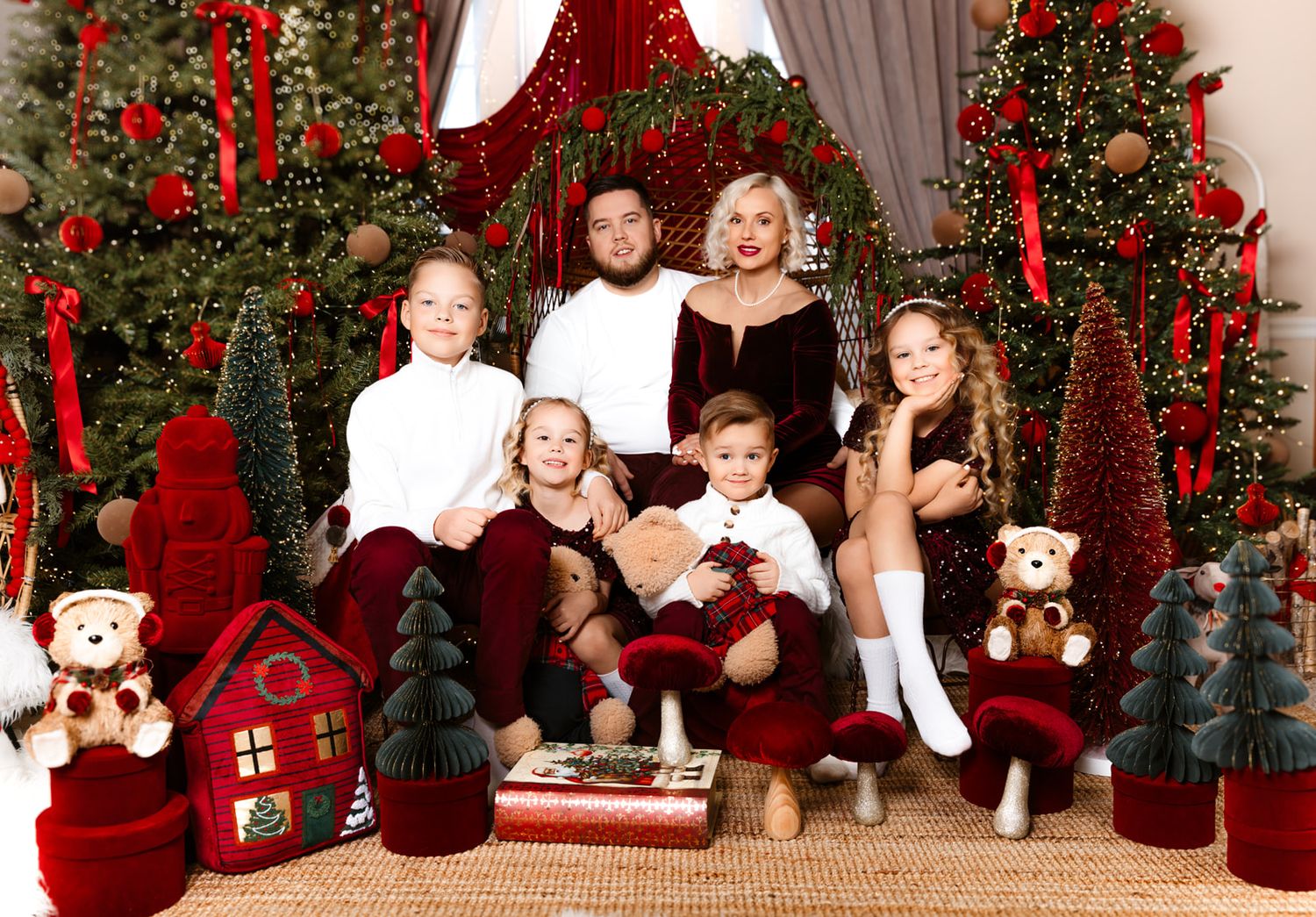 Family Christmas portrait with parents and four children posed among festive red and green holiday decorations and trees.