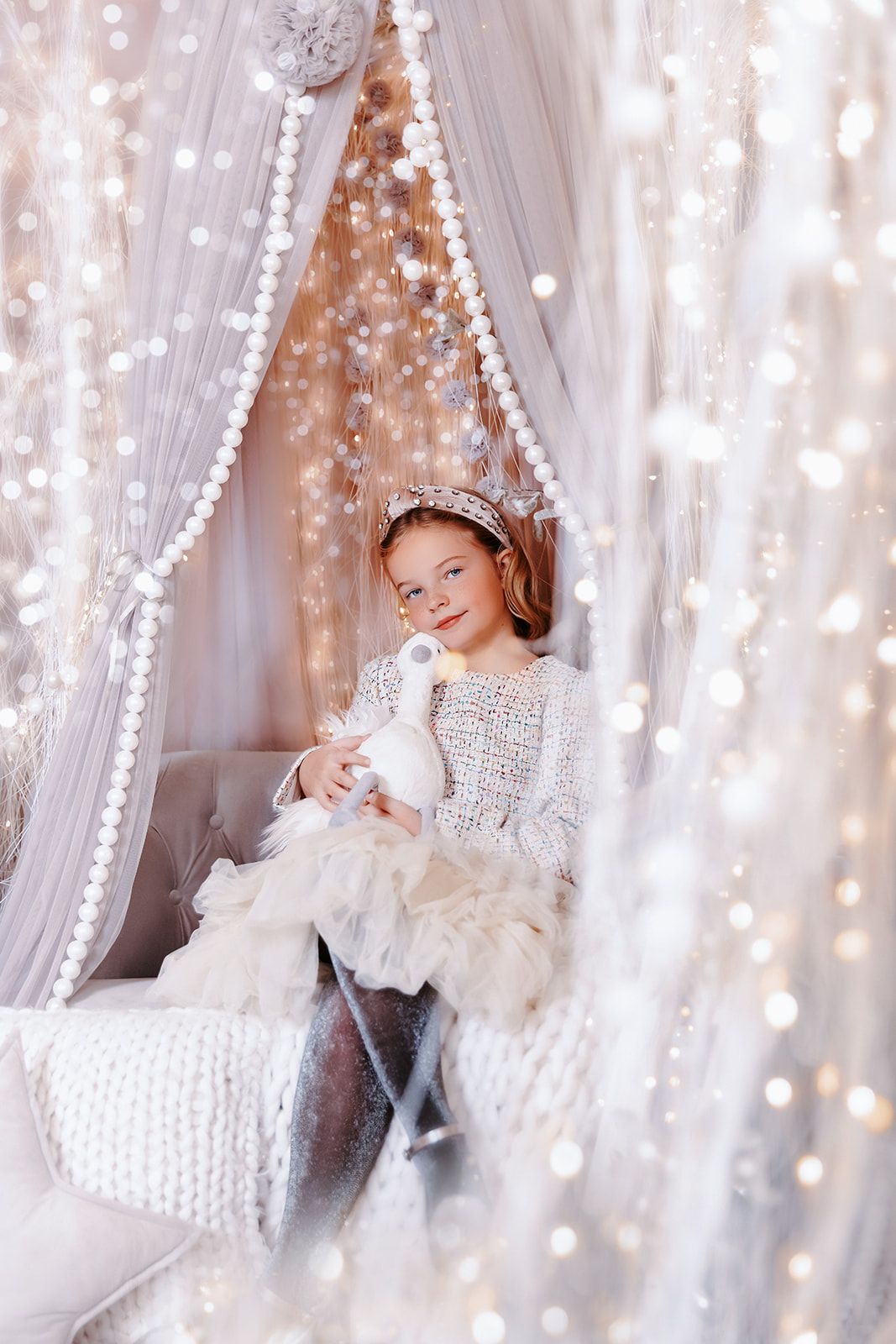 Young girl in white dress holding a toy unicorn, sitting in a fairy-tale canopy bed with sparkling lights.