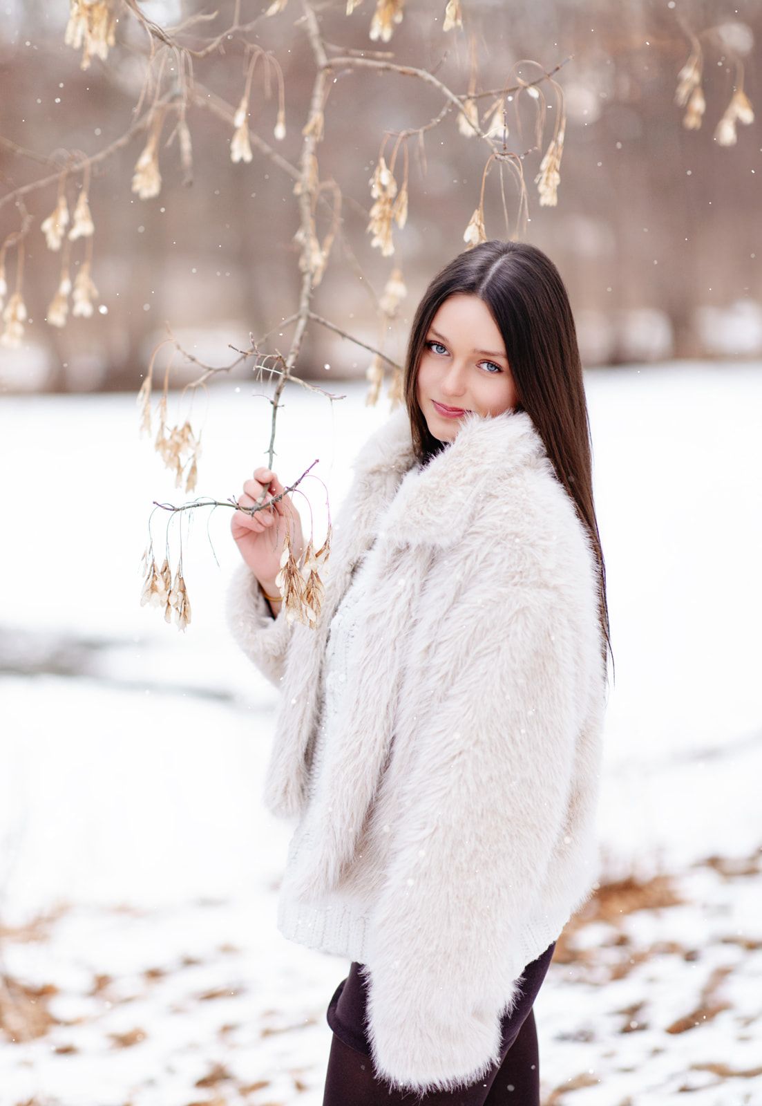 Young woman in white fur coat holding a bare branch in a snowy winter park, looking over her shoulder.