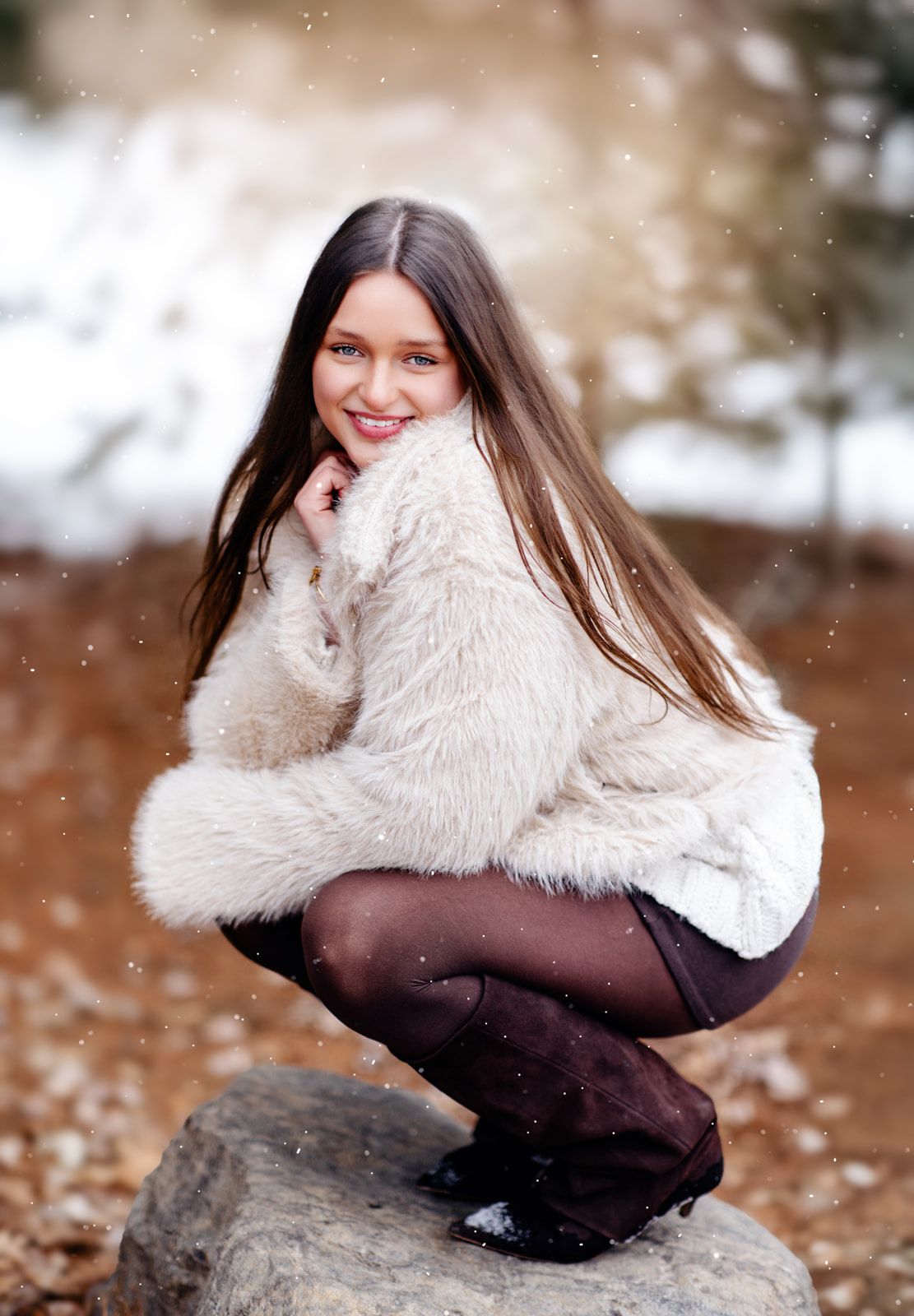 Smiling young woman in cream faux fur coat crouching on rock in snowy winter setting.