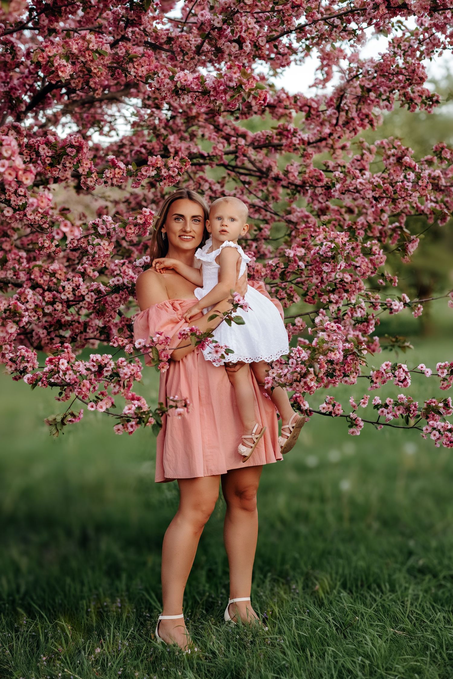 Beautiful mother in pink off-shoulder dress holding baby in white dress by pink blooming tree.
