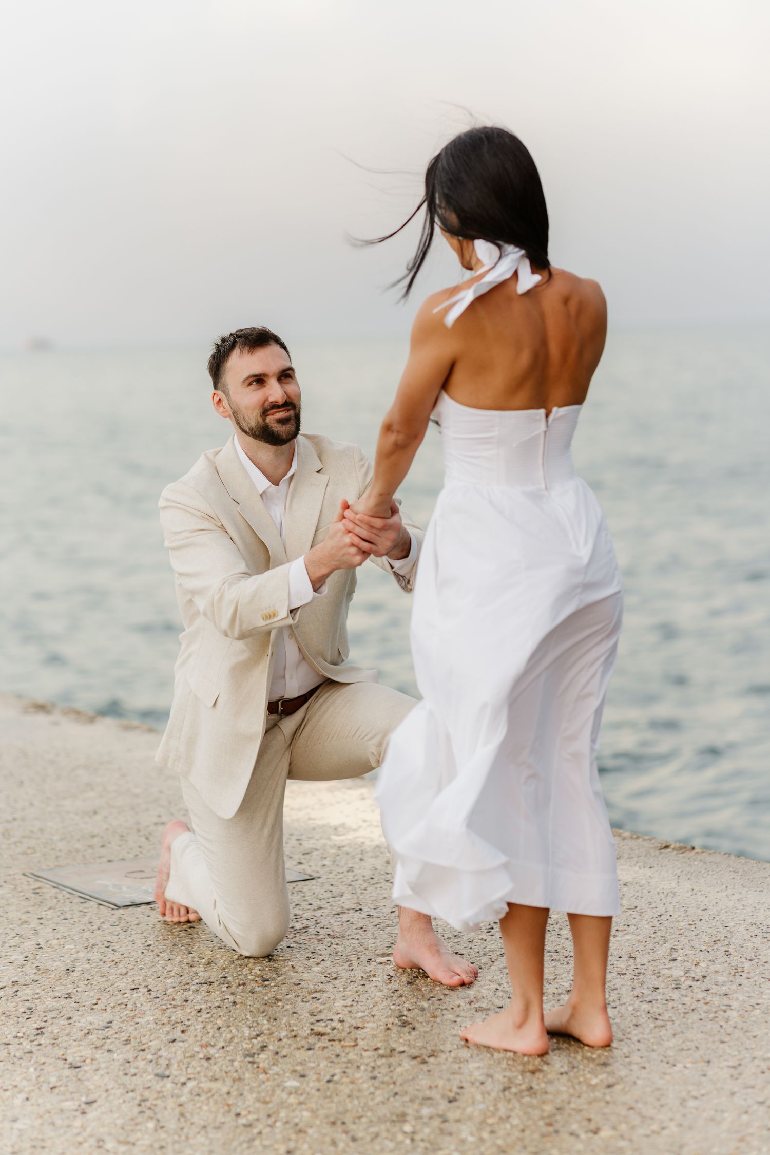 Smiling man on one knee holding woman's hands during beach proposal with ocean backdrop.