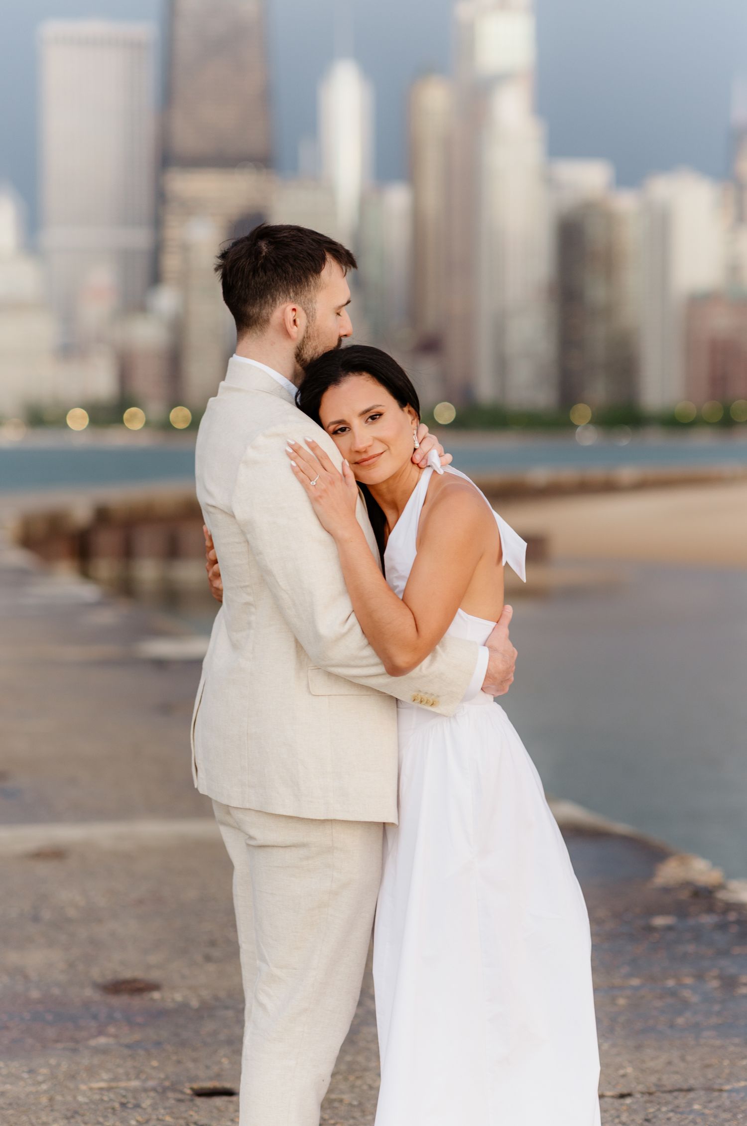 Couple embracing on Chicago lakefront, bride in white dress and groom in beige suit with city skyline backdrop.