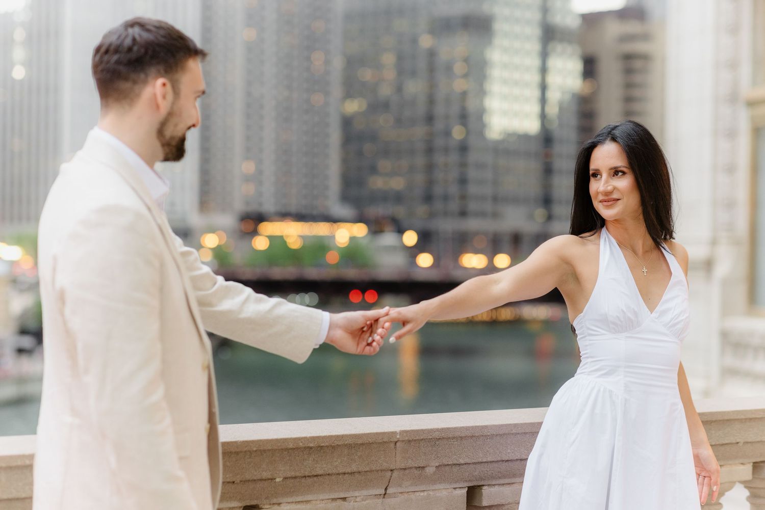 Couple holding hands on a Chicago riverwalk bridge, woman in white dress smiling at camera with city skyline behind.