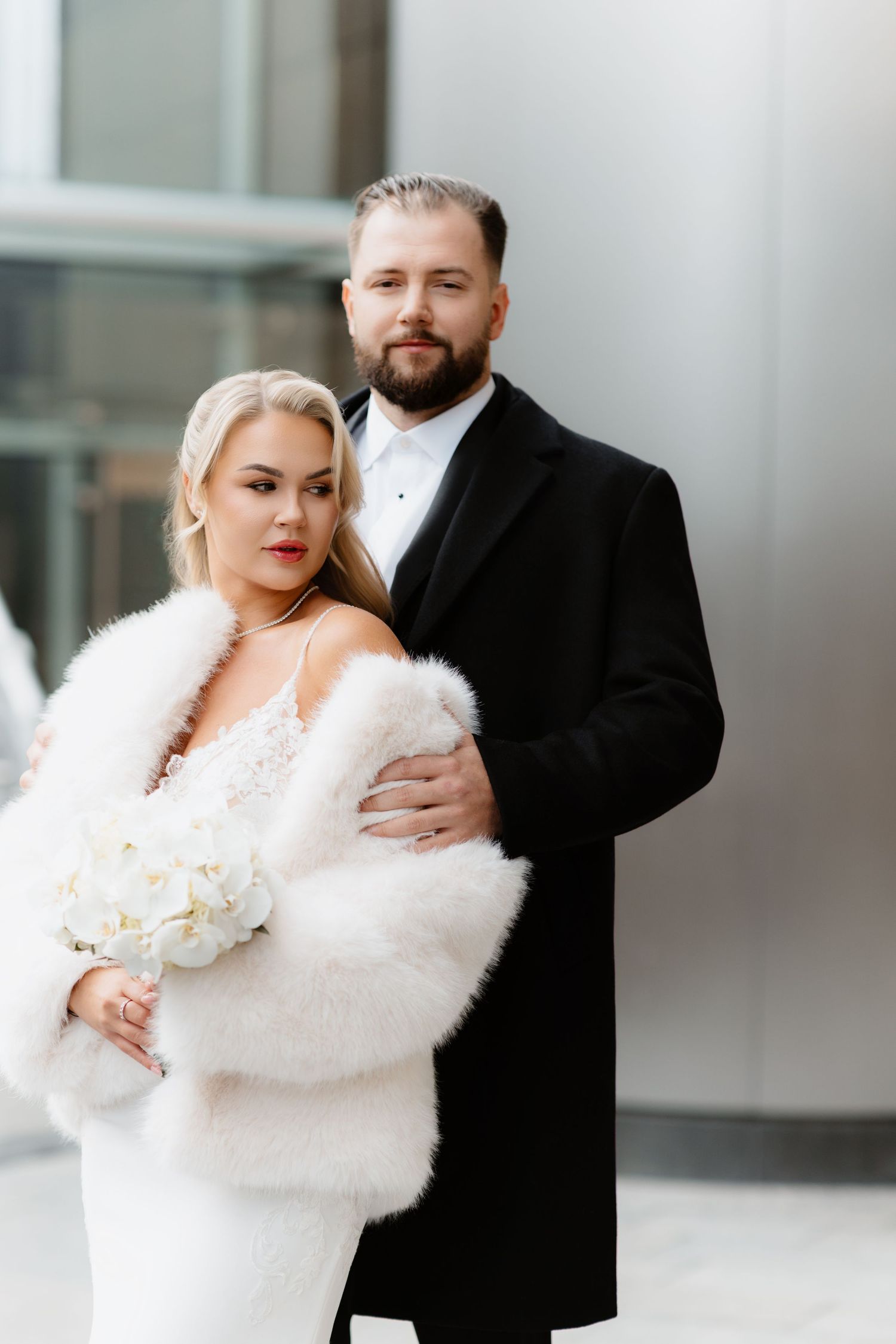 Bride in white fur wrap holds bouquet as groom embraces her in Chicago.