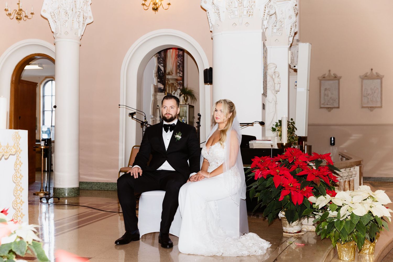 Newlyweds sitting together at church altar surrounded by festive red poinsettia floral arrangements.