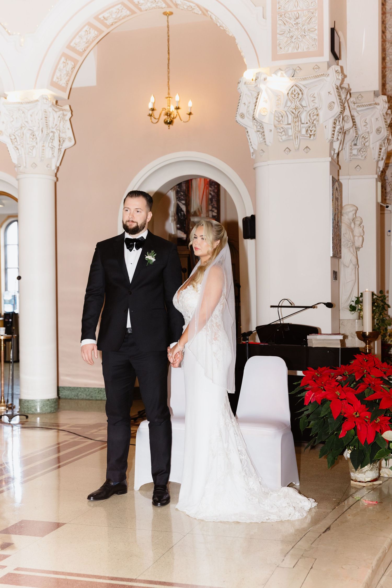 Bride in white lace gown and groom in black tuxedo standing in ornate church archway.