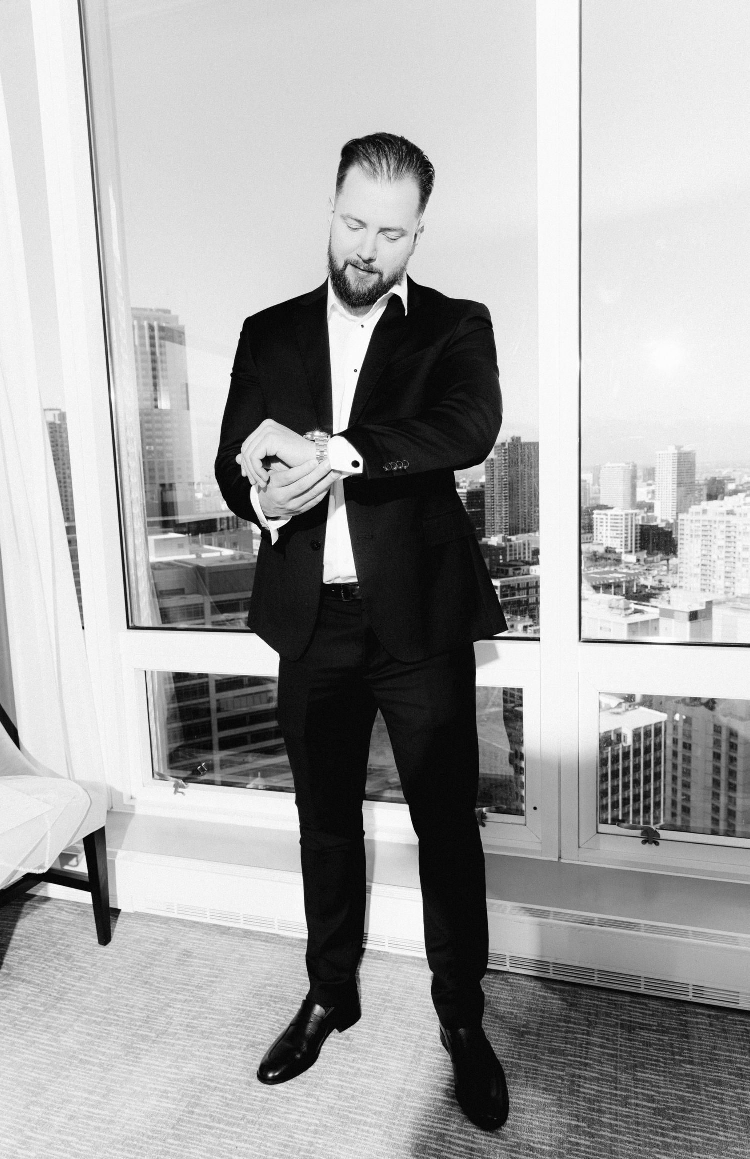 Black and white photo of bearded groom in black suit checking his watch by high-rise windows with city skyline view.