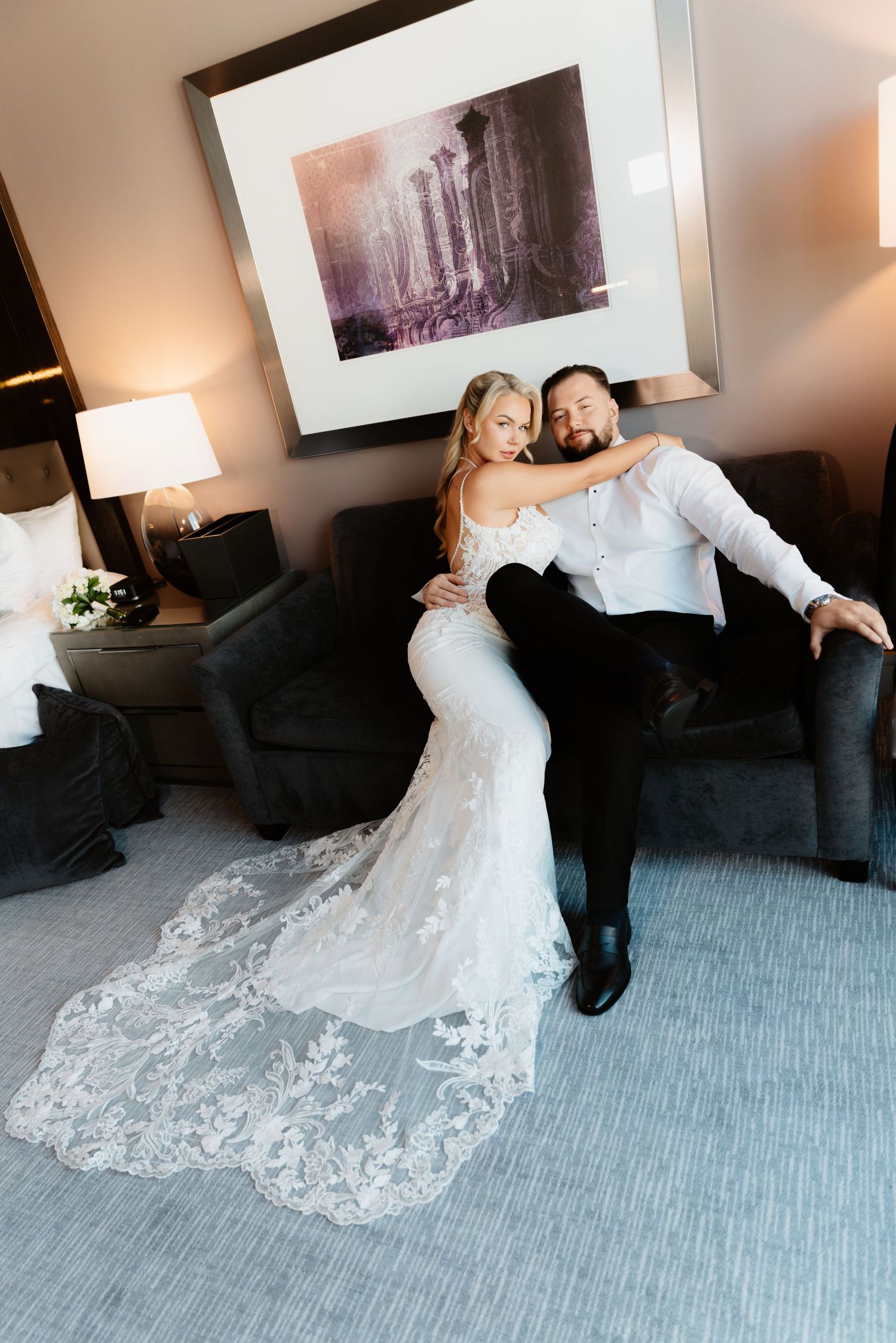 Bride in lace wedding gown sits with groom in white shirt on hotel room sofa, color photo.